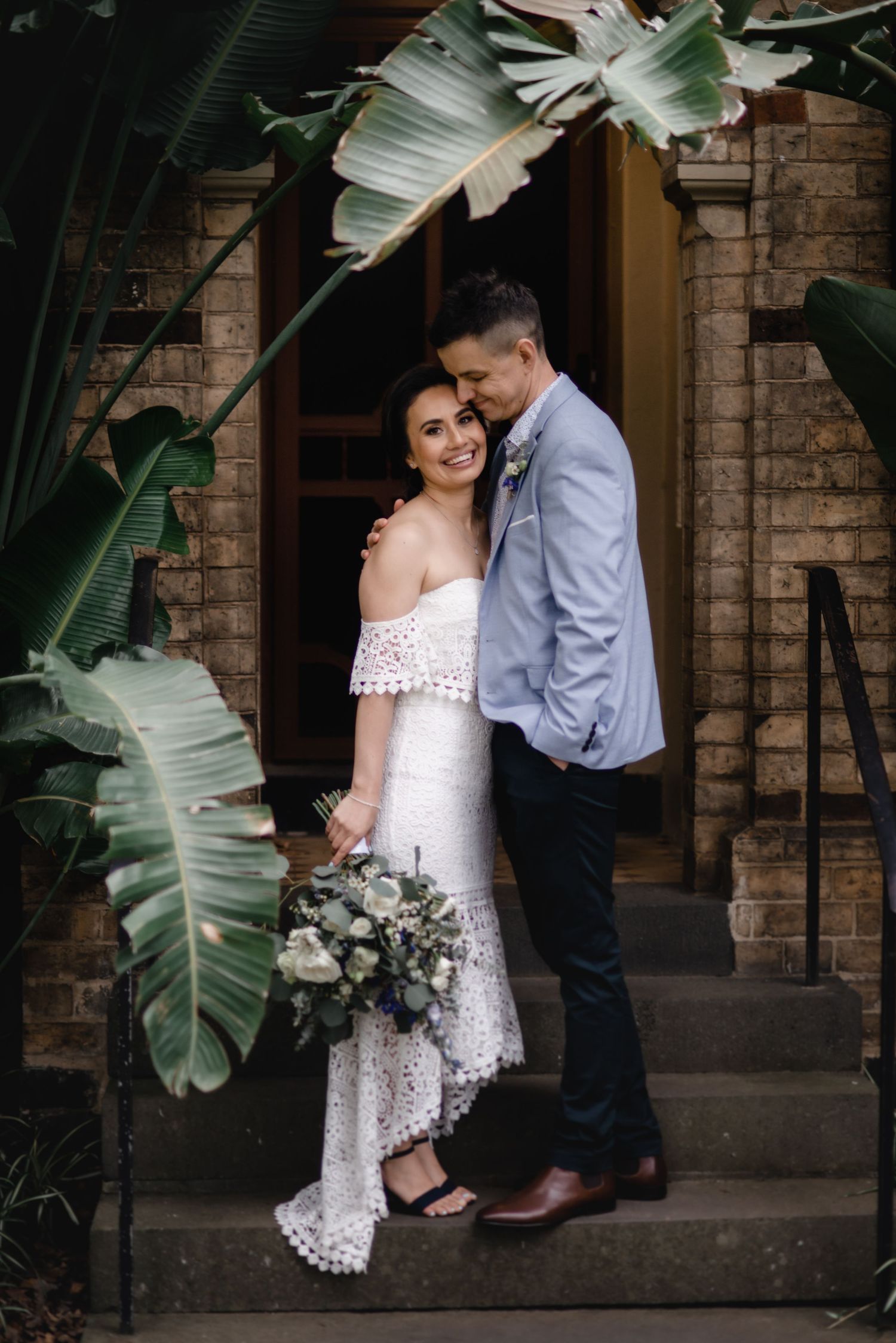 A romantic wedding portrait framed by tropical leaves against a brick entrance.