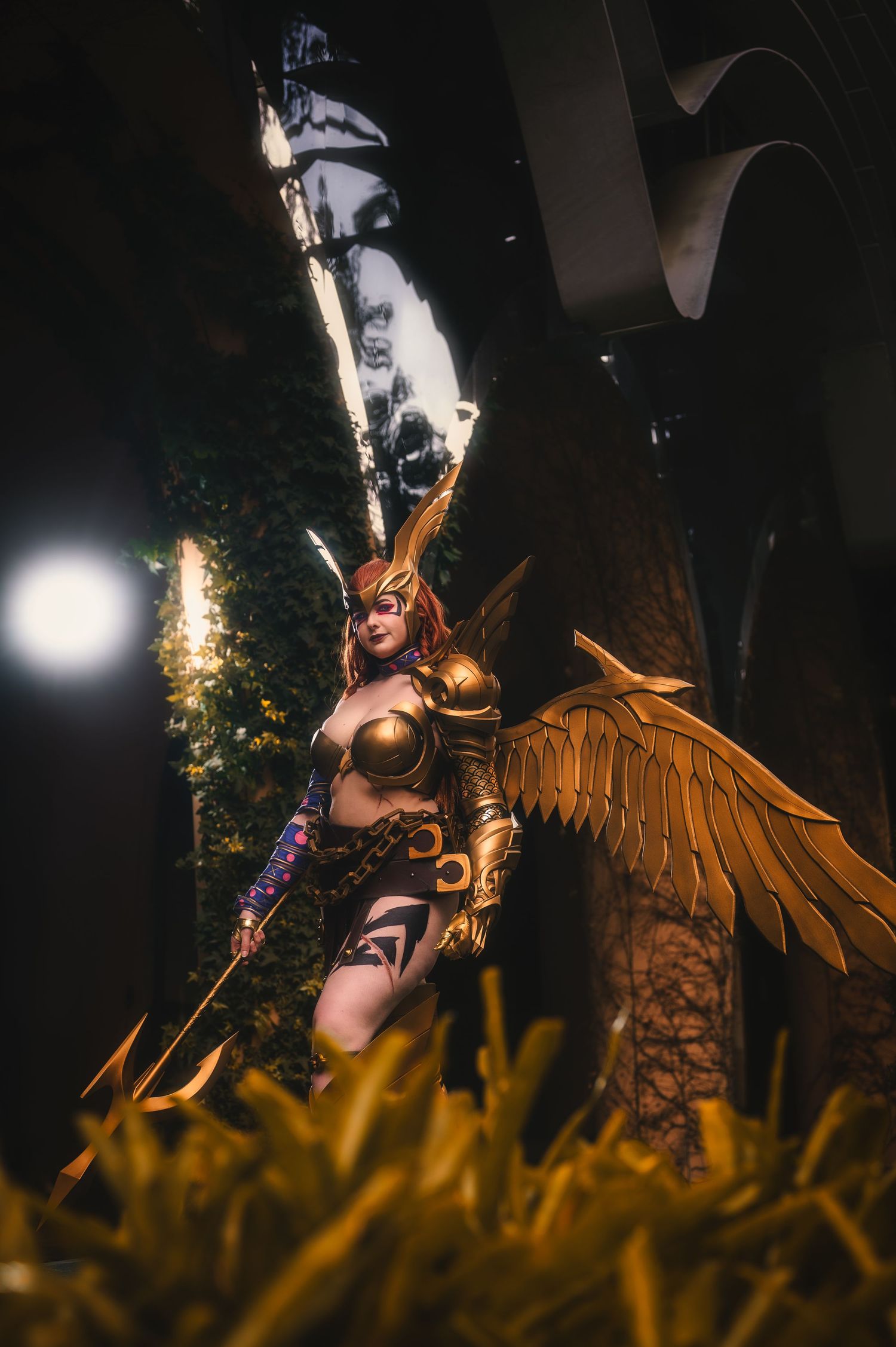 Woman cosplaying as Hawkgirl in golden armor with wings and spear, posed dramatically outdoors at night.