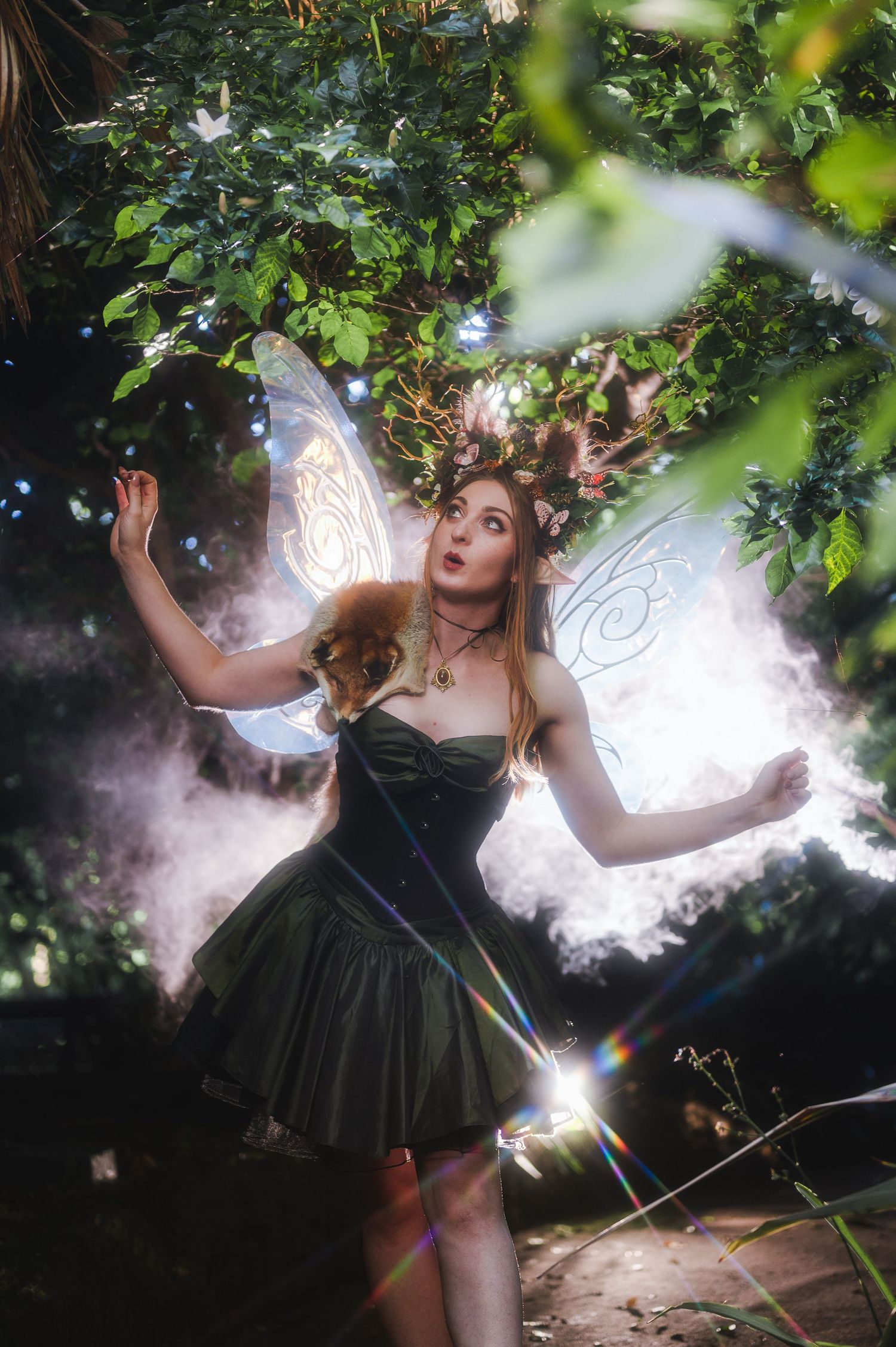 A woman in a green fairy costume with wings and floral headpiece poses in a misty garden.