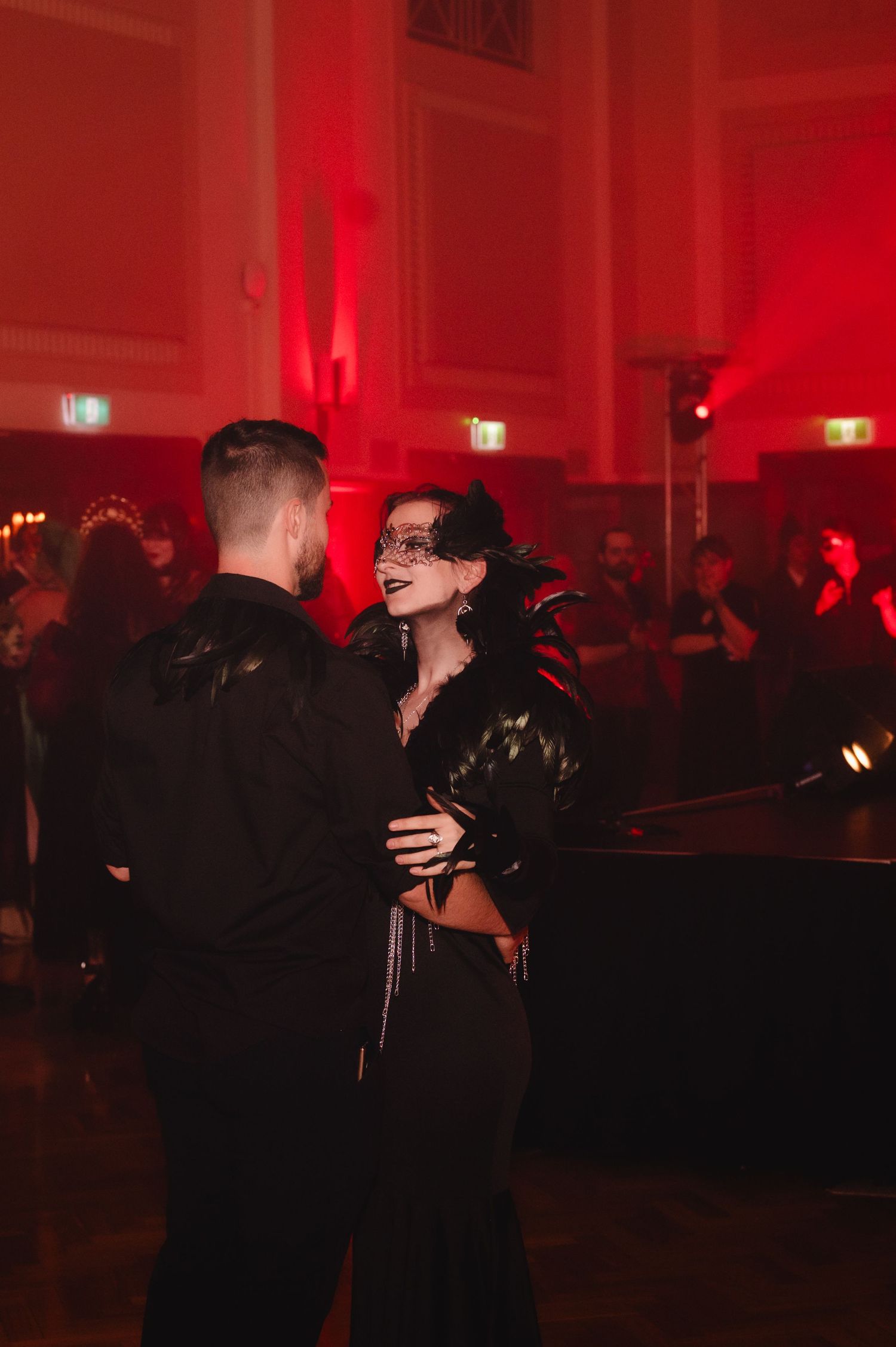 Couple dancing at a red-lit masquerade ball, woman in black outfit with feathered mask, elegant gothic atmosphere.