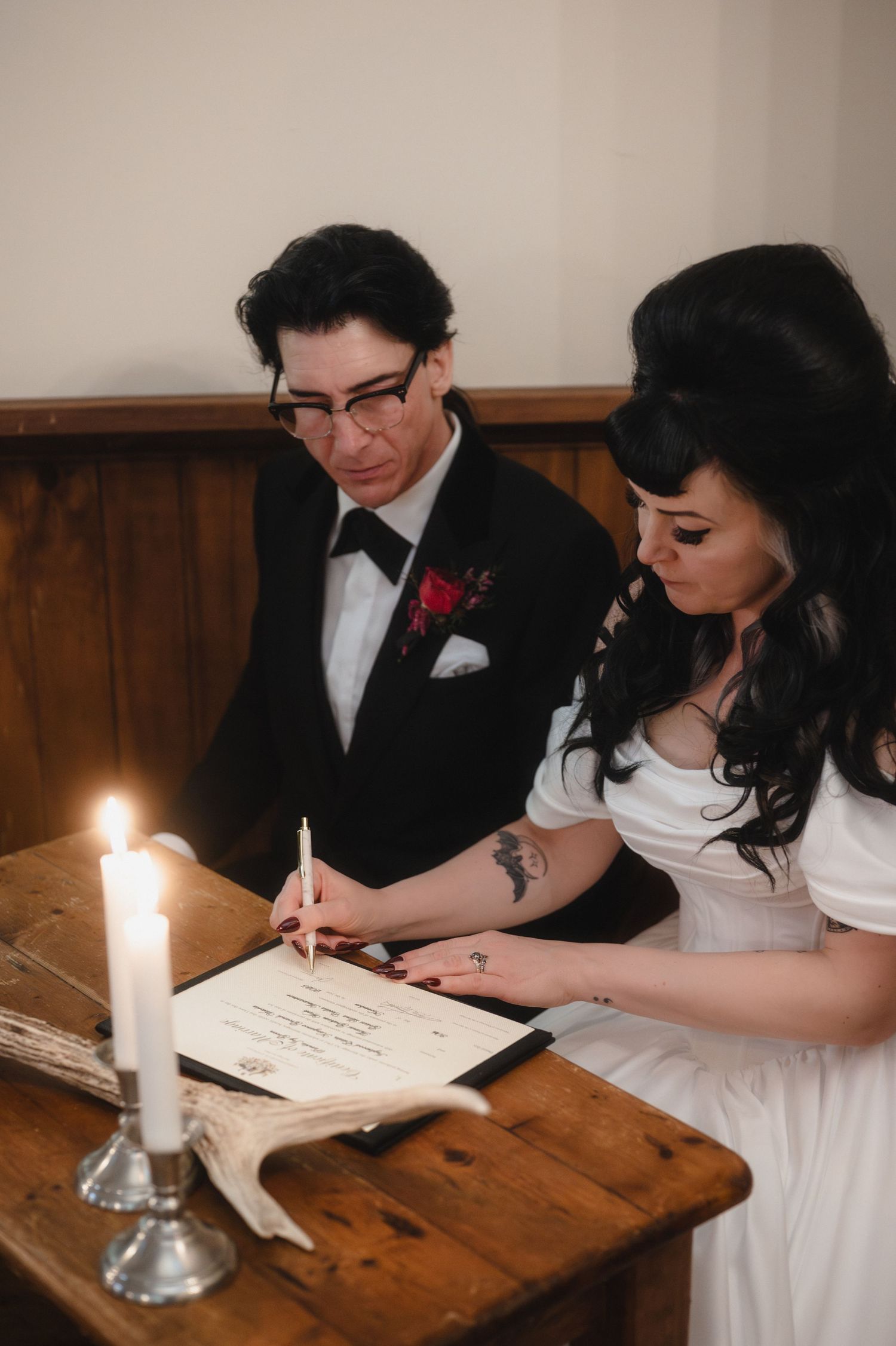 Couple signing marriage documents at a rustic wooden table with a lit candle in an intimate ceremony setting.