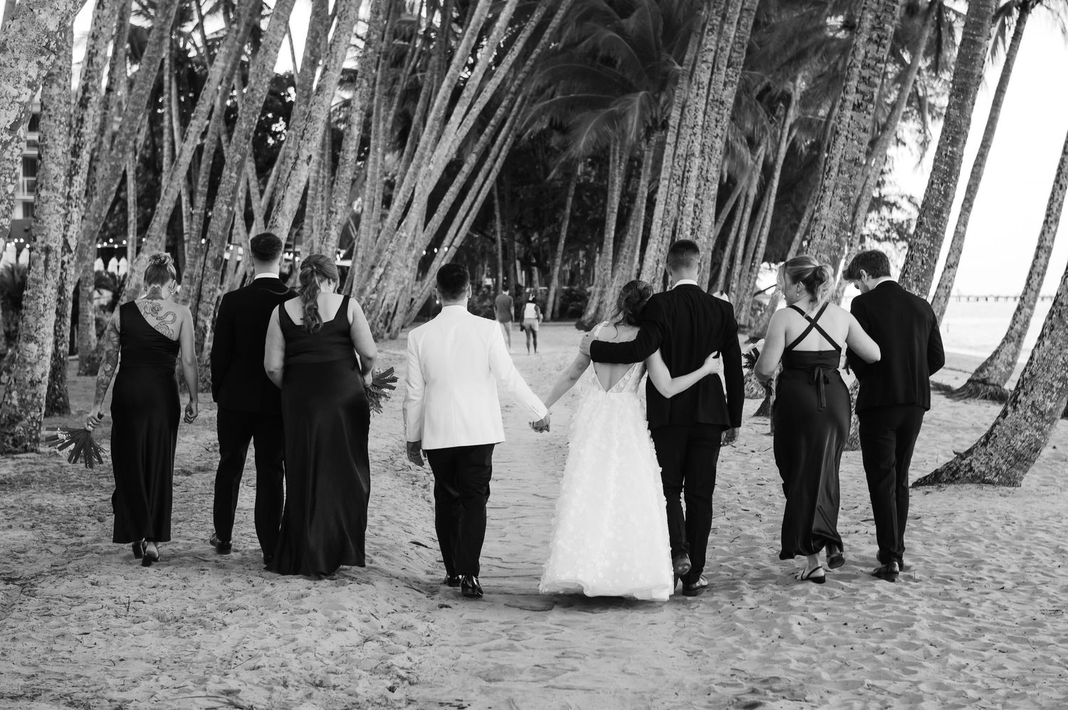 Wedding party poses in black and white formal attire on sandy beach with trees.