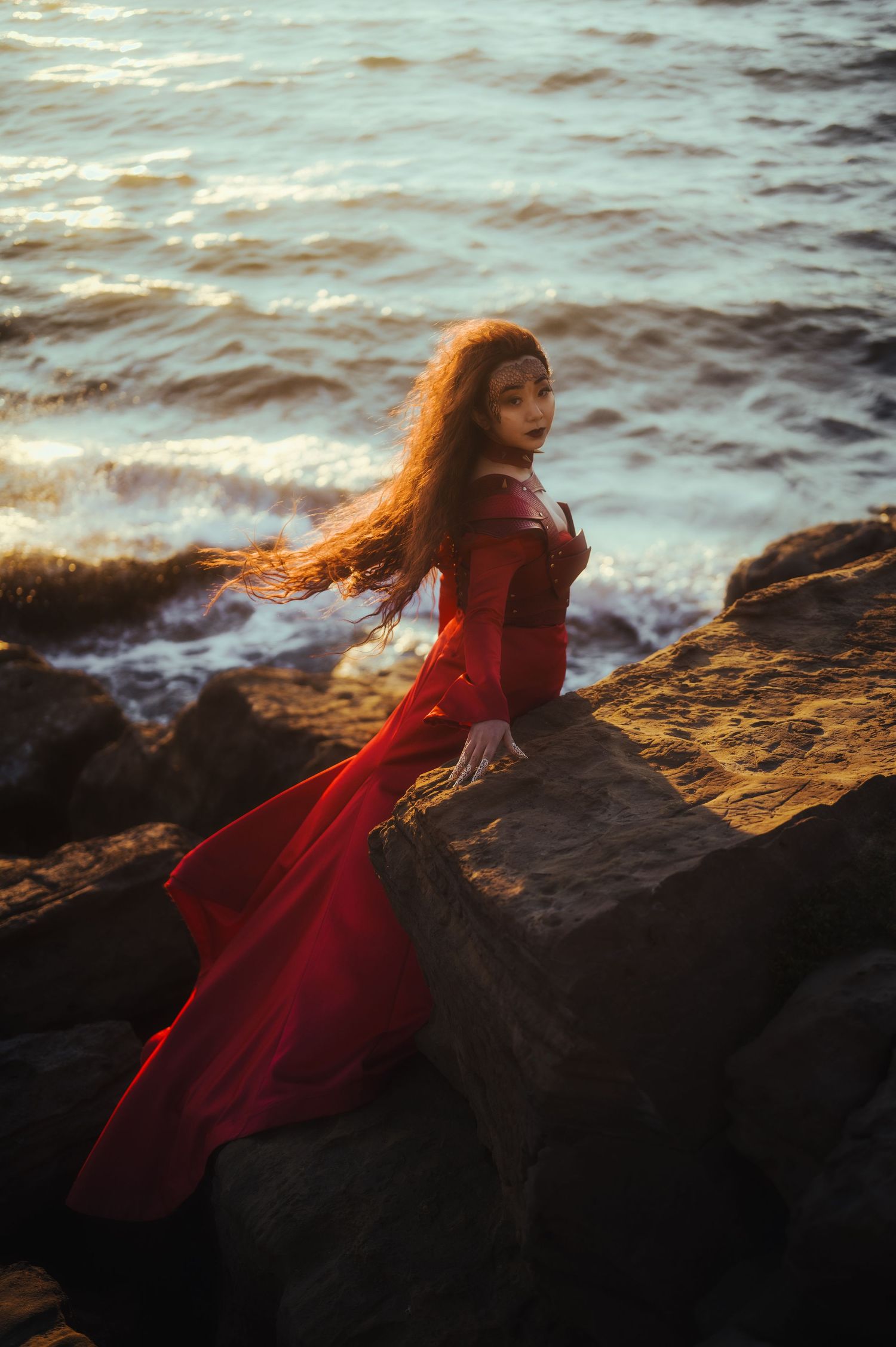 A flowing red dress catches the wind as a figure poses dramatically on coastal rocks at sunset.