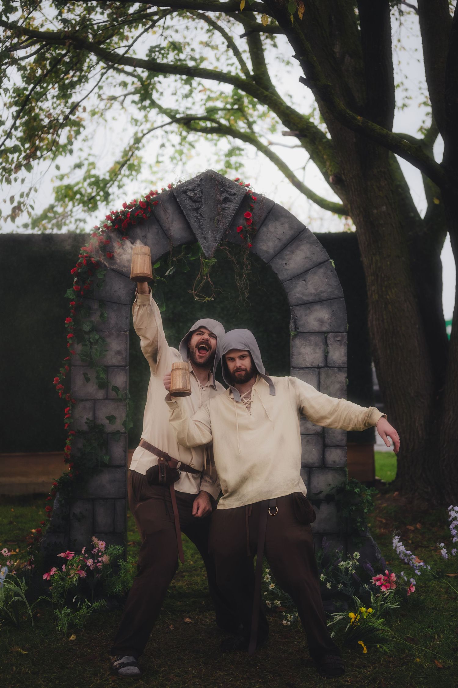 Two men in medieval costumes raising cups under a stone arch decorated with flowers and greenery outdoors.