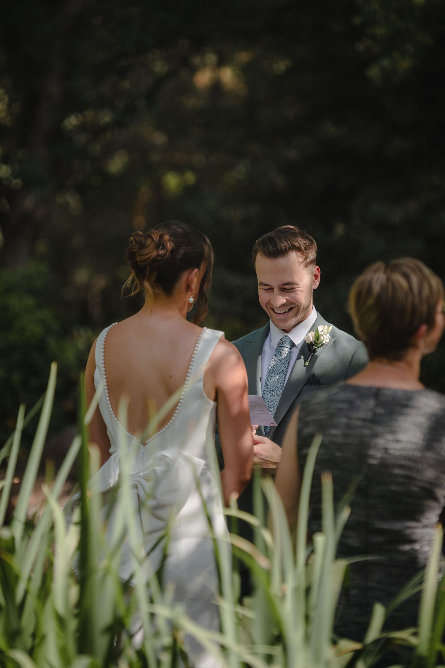 Groom smiling joyfully at bride during outdoor garden wedding ceremony surrounded by lush greenery.