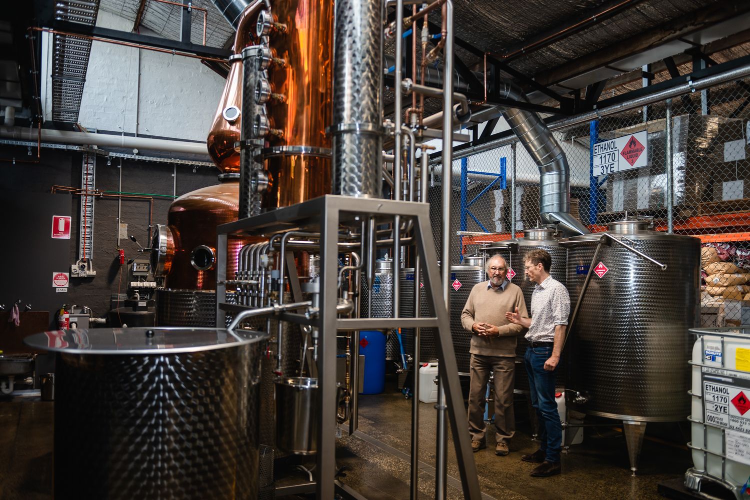 Two men conversing beside a copper distillery still and fermentation tanks in an industrial craft spirits distillery.