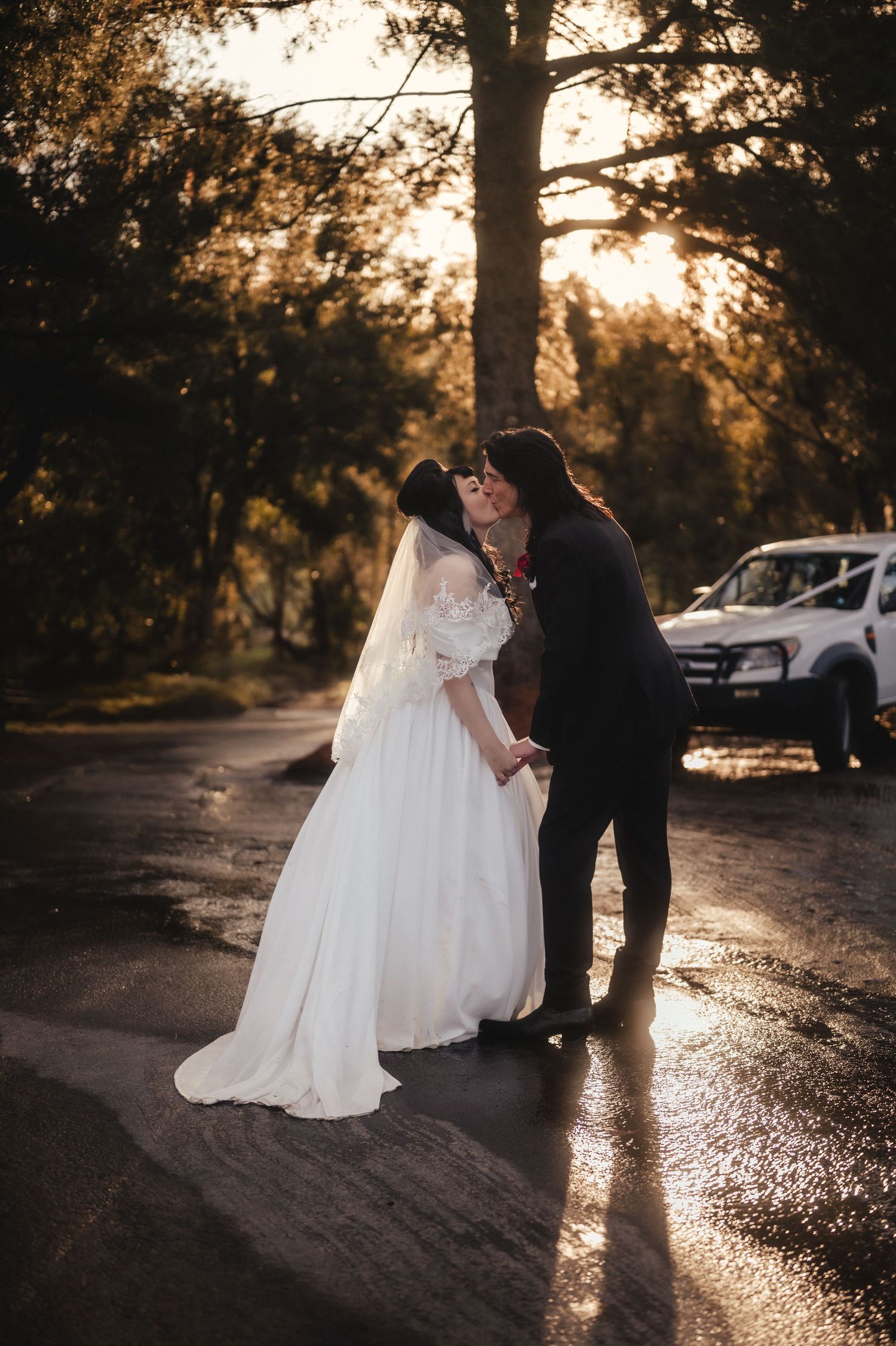 Newlyweds share a romantic kiss on a wet road bathed in warm golden sunset light.