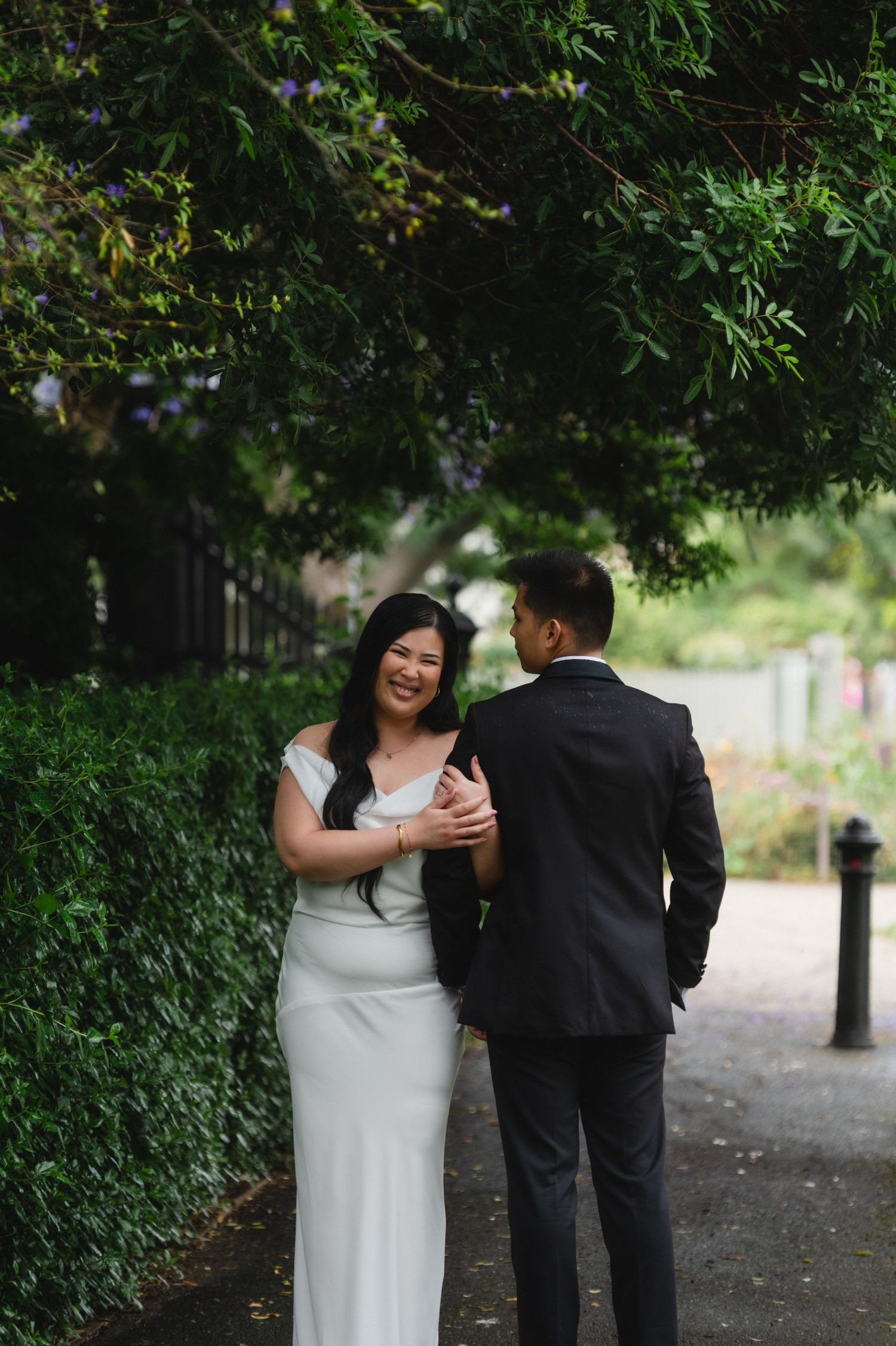 A smiling bride in a white off-shoulder gown holds hands with a groom in a dark suit on a tree-lined path.