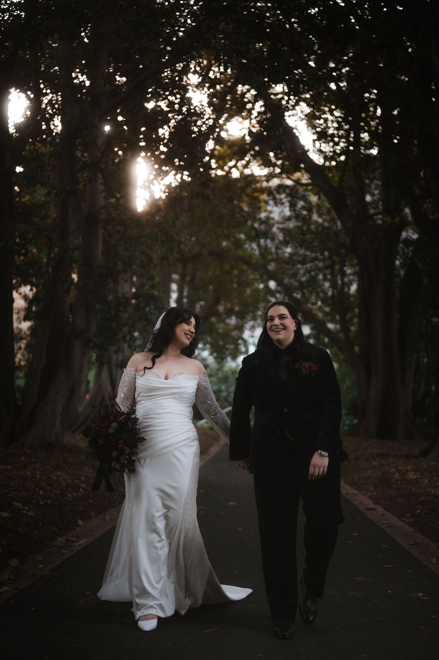 A couple in wedding attire walks along a tree-lined path at dusk creating a romantic silhouette.