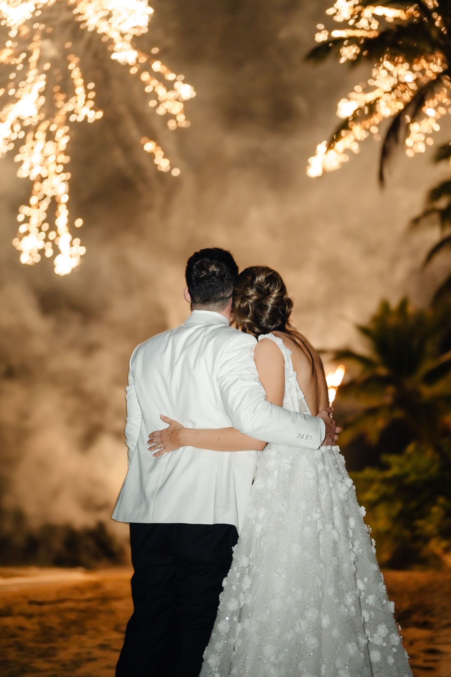 Couple embracing under twinkling lights during their evening beach wedding celebration.