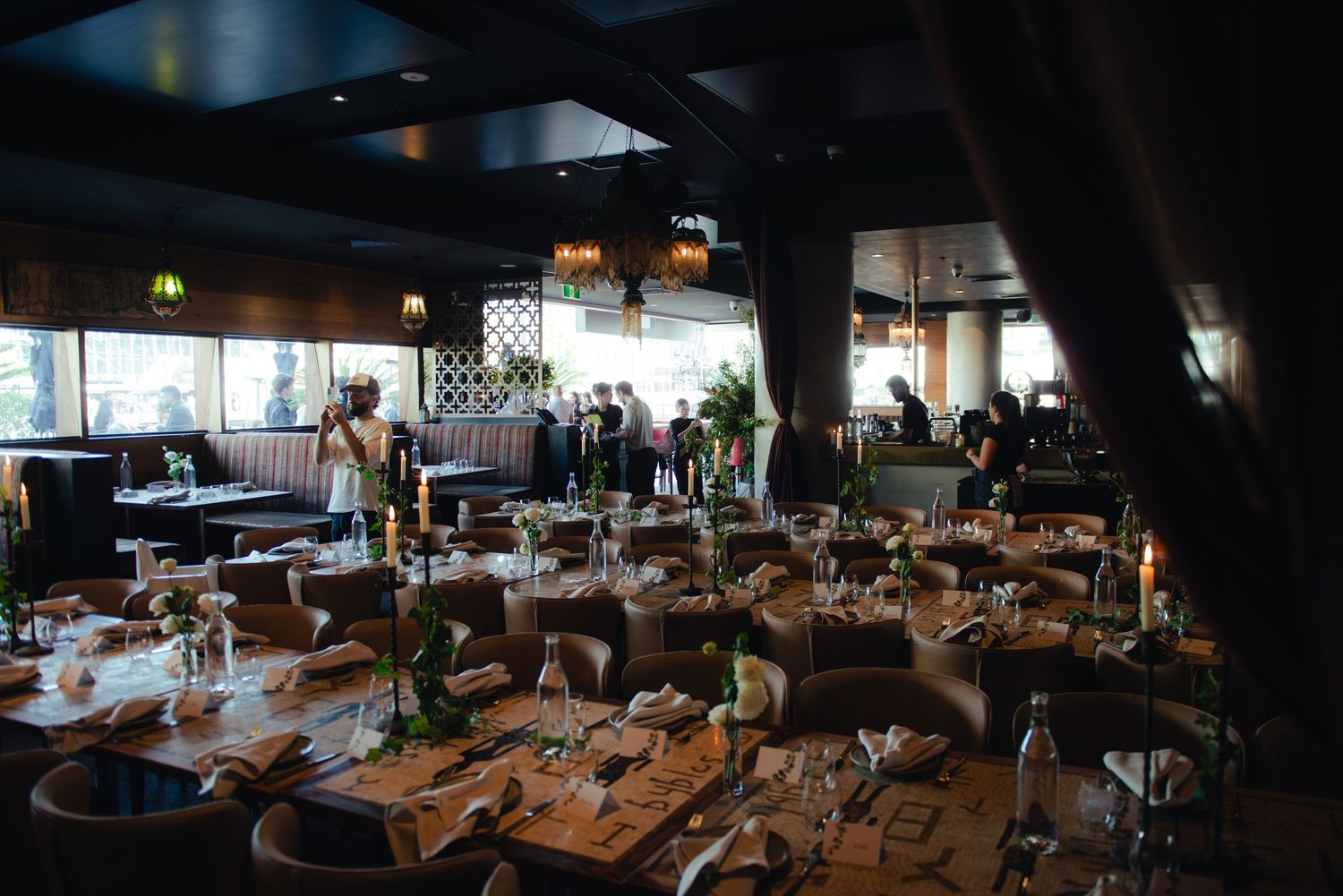Elegant restaurant dining room with candlelit tables set for a formal event, featuring warm lighting and guests mingling in background.