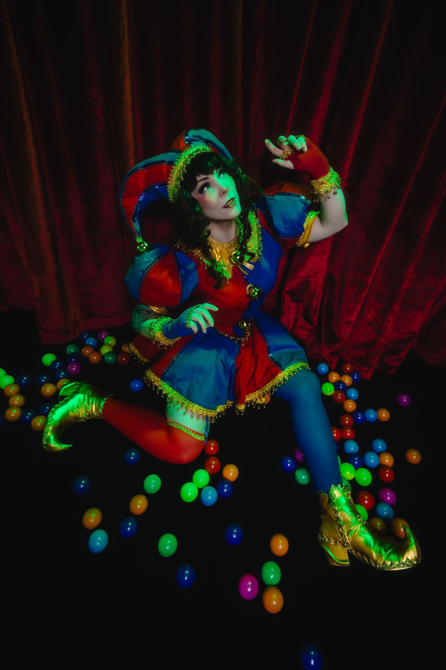 Colorful jester costume cosplay with glowing balls, red curtain backdrop, dramatic pose under UV light.