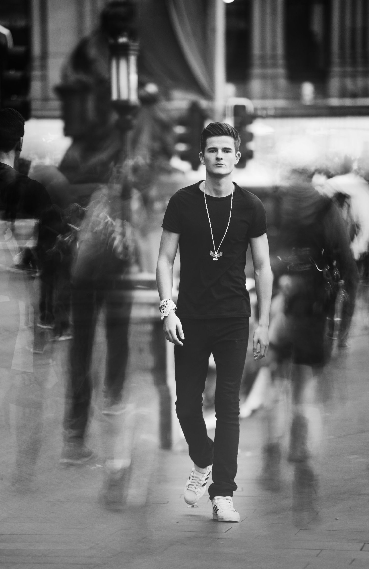 Young man in black t-shirt and jeans walks confidently through a busy public space, captured in black and white.