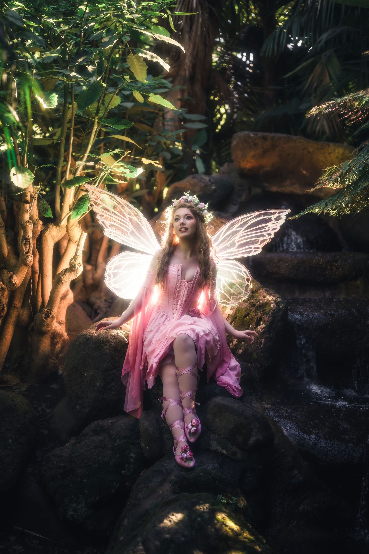 A woman in a pink fairy costume with glowing wings and floral crown sits on rocks in a lush forest setting.