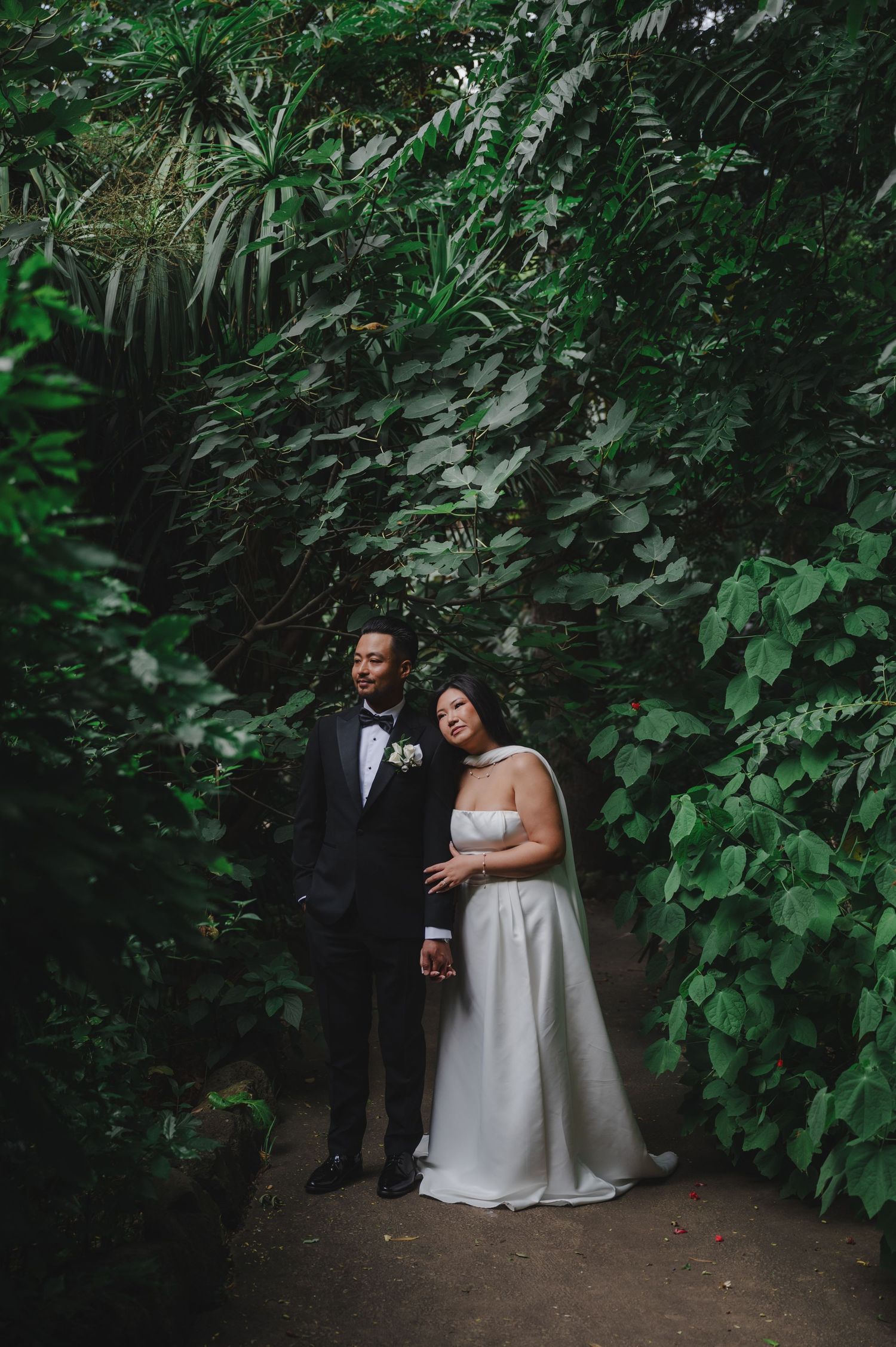 Bride and groom pose among lush tropical greenery in a romantic garden wedding portrait.