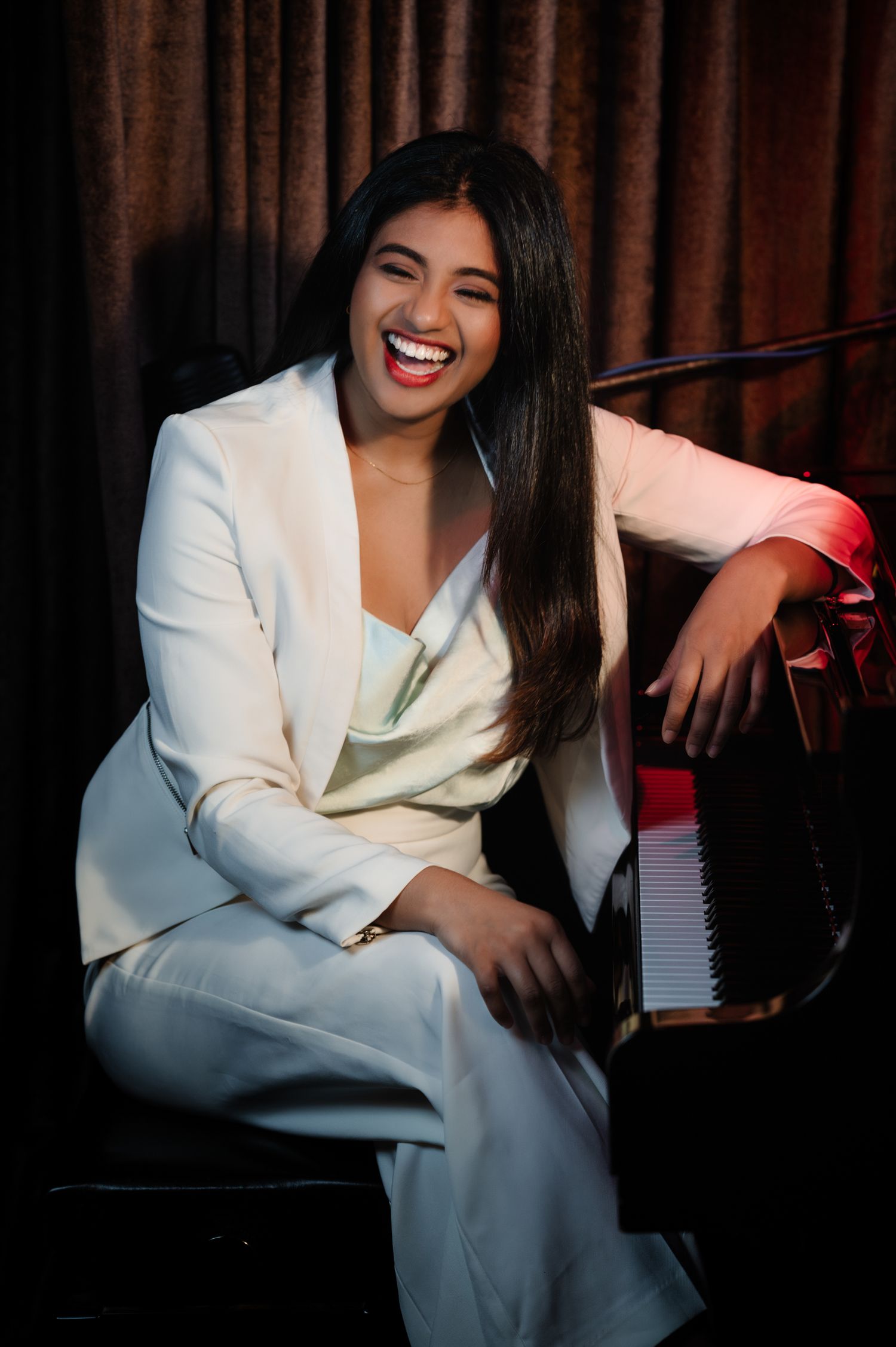 A performer in a white blazer laughs joyfully while seated at a piano in a dimly lit venue.