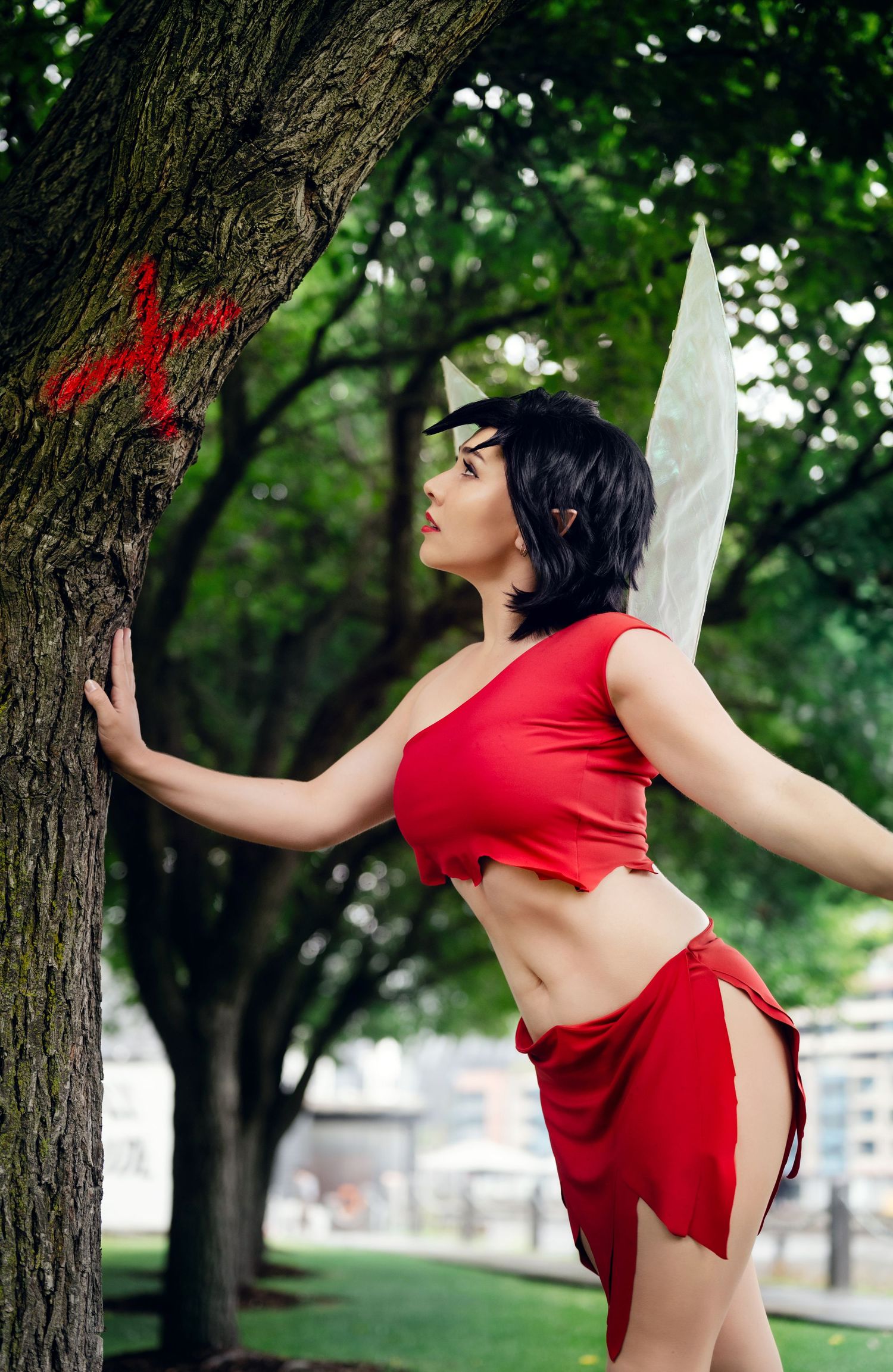 Fantasy-inspired cosplay in a red outfit posed against a tree in a park setting.