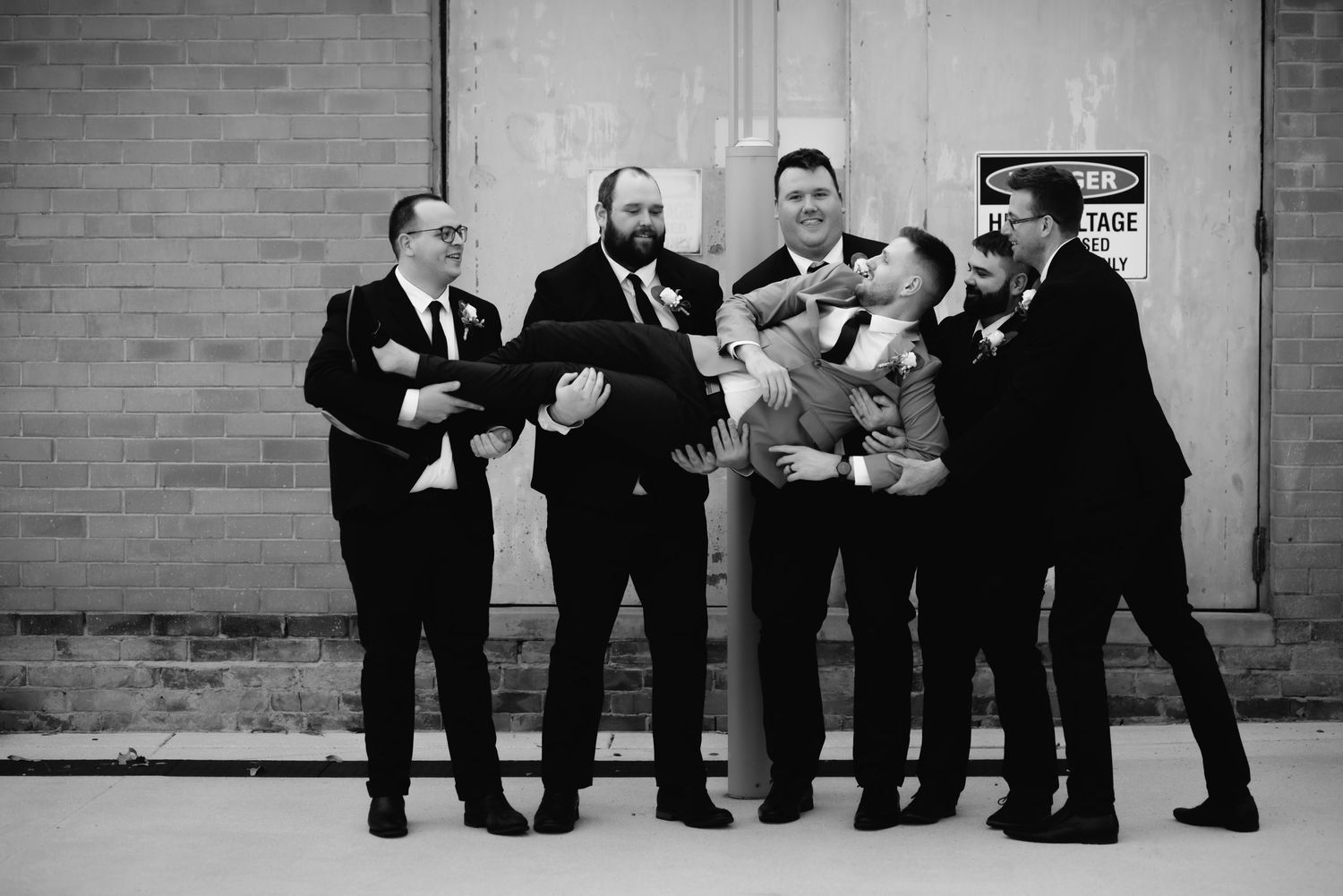 Groomsmen pose casually against brick wall in black and white photograph.