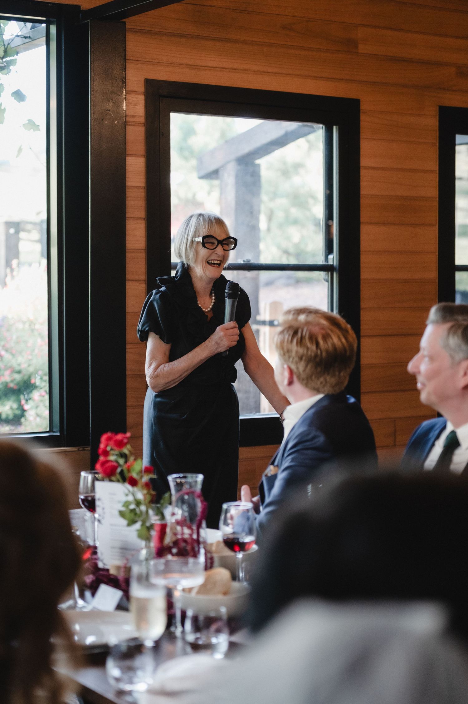 Elegant older woman with gray hair gives a heartfelt wedding speech to seated dinner guests.
