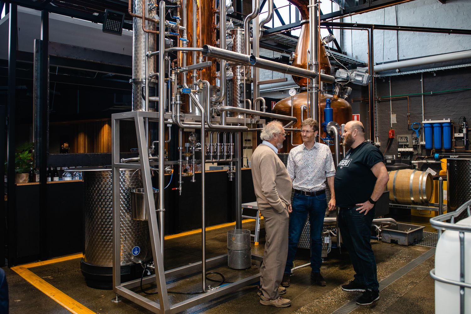 Three men conversing beside copper pot stills and distillery equipment in a craft spirits distillery.
