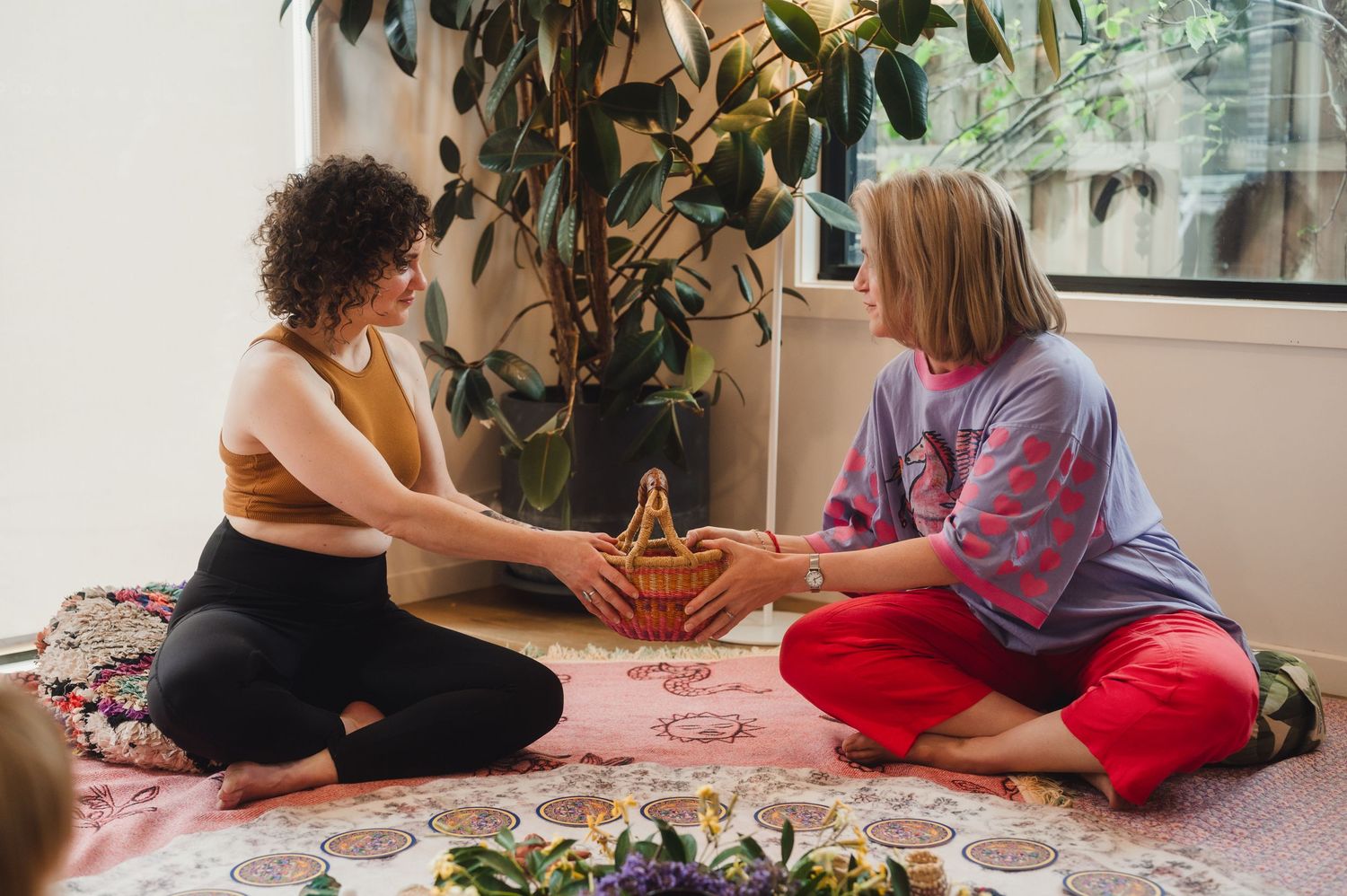 Two women sitting cross-legged on a floral mat, sharing a woven basket in a plant-filled room.