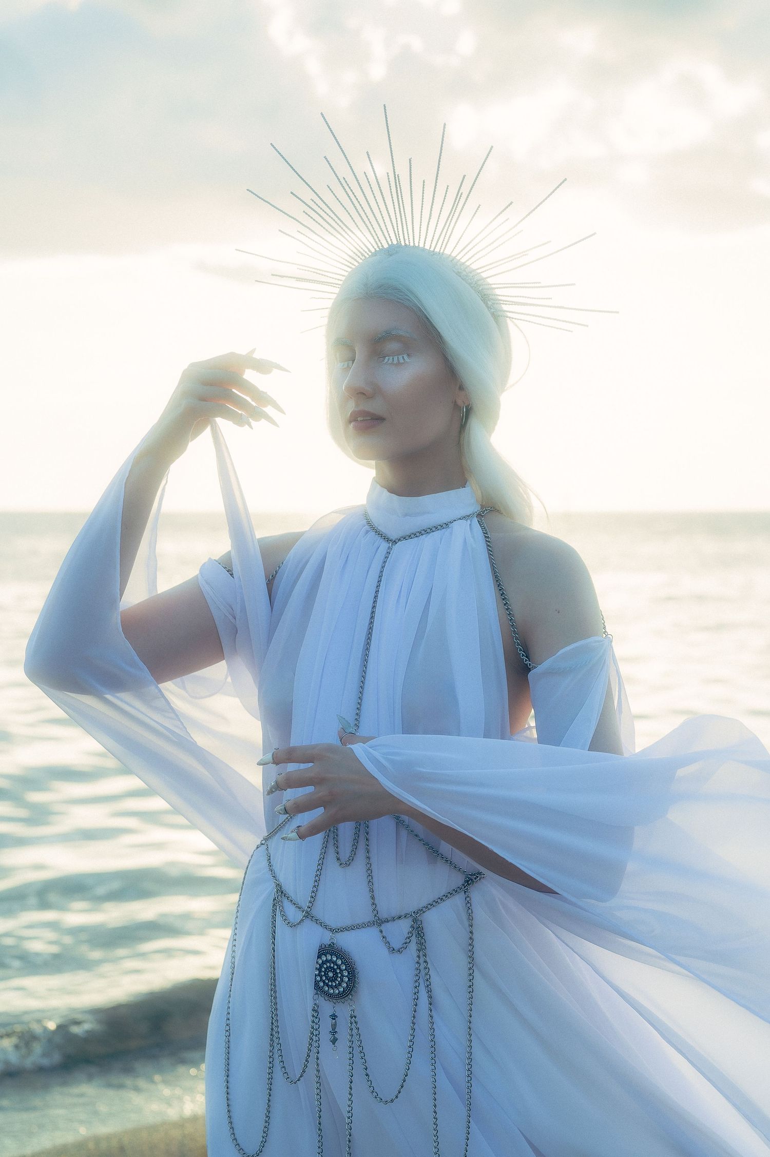 Ethereal figure in flowing white dress and spiked crown poses against ocean backdrop in dreamy sunlit scene.