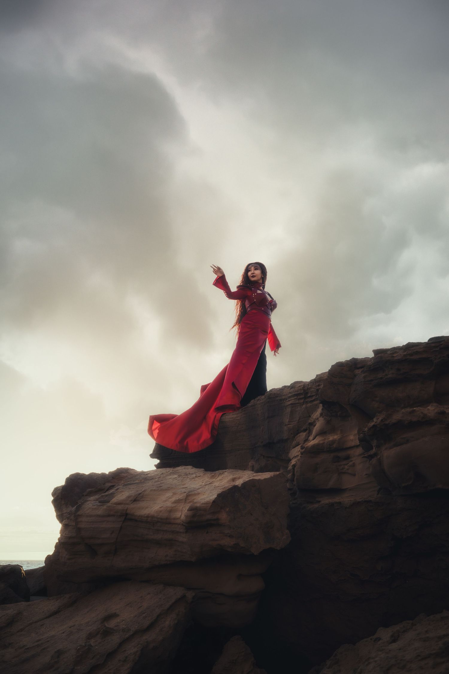 A silhouette in a flowing red dress stands atop dramatic rock formations against a cloudy sky.