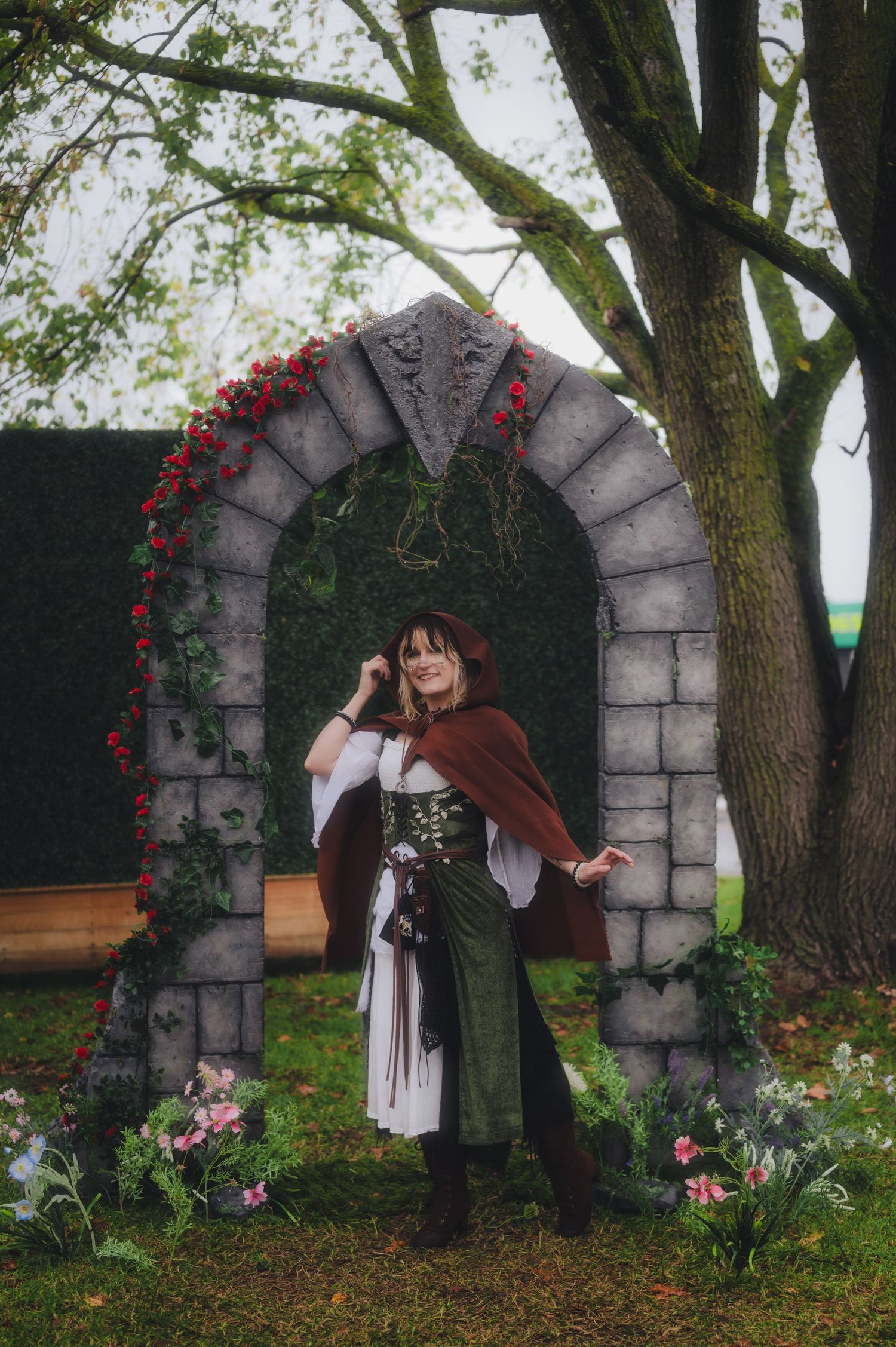 Woman in medieval fantasy costume poses under a stone arch decorated with red roses and greenery outdoors.