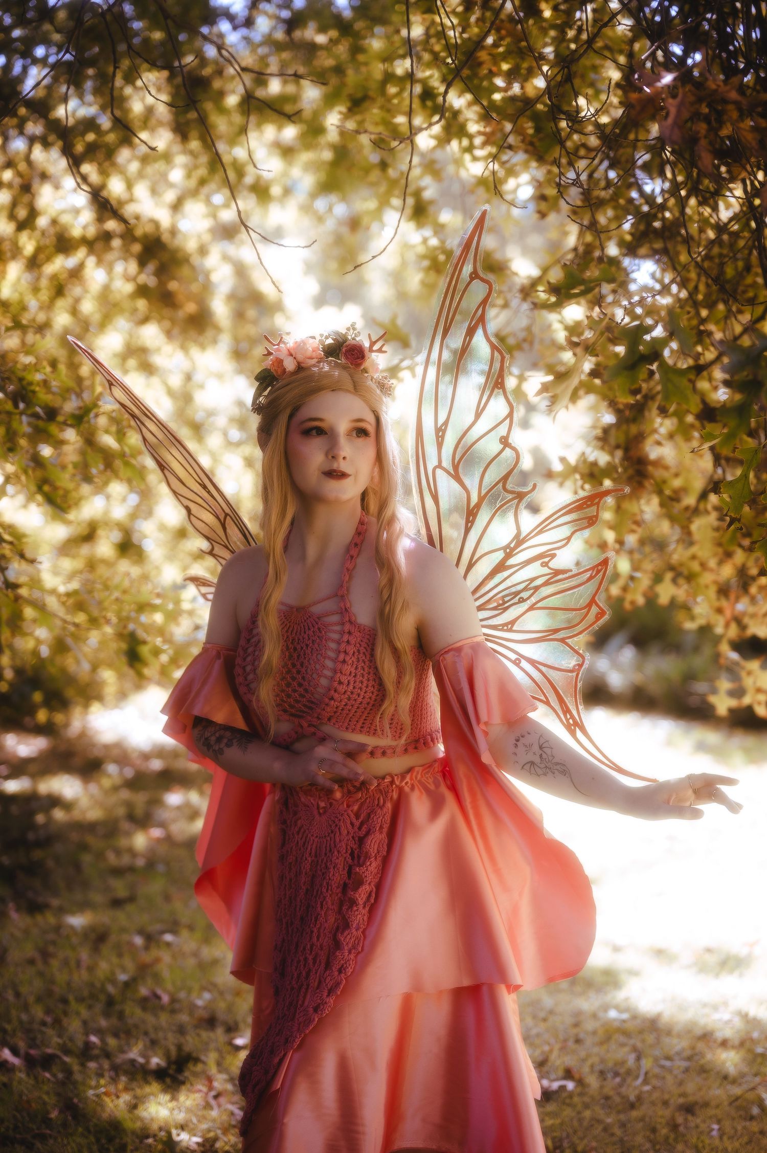 A woman in a pink fairy costume with translucent wings and floral crown poses in a sunlit forest.