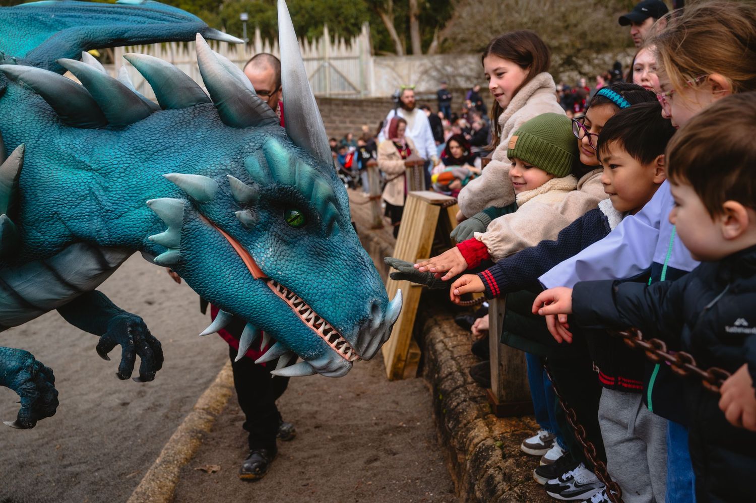 Children reach out to touch a realistic teal dragon costume at an outdoor event, with a crowd of excited spectators behind them.
