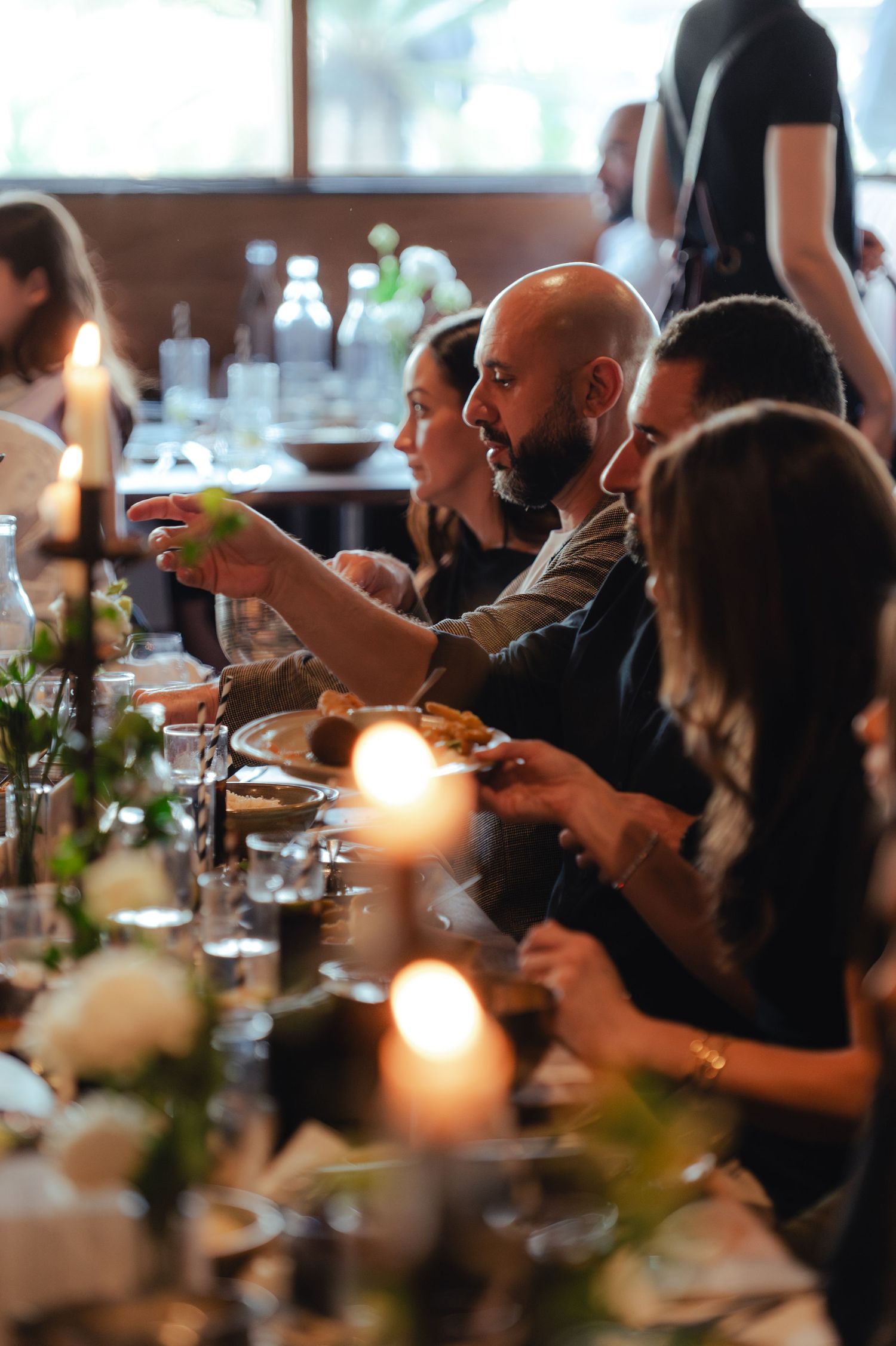 Guests dining at a candlelit dinner party, with a bald man reaching across the table.
