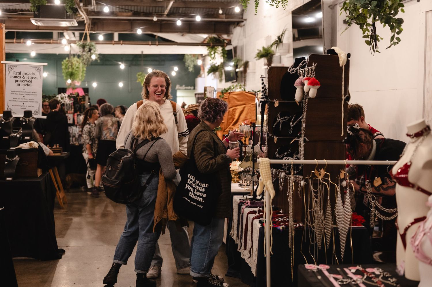 Shoppers browse colorful clothing racks at a busy indoor vintage market with hanging plants and warm lighting.