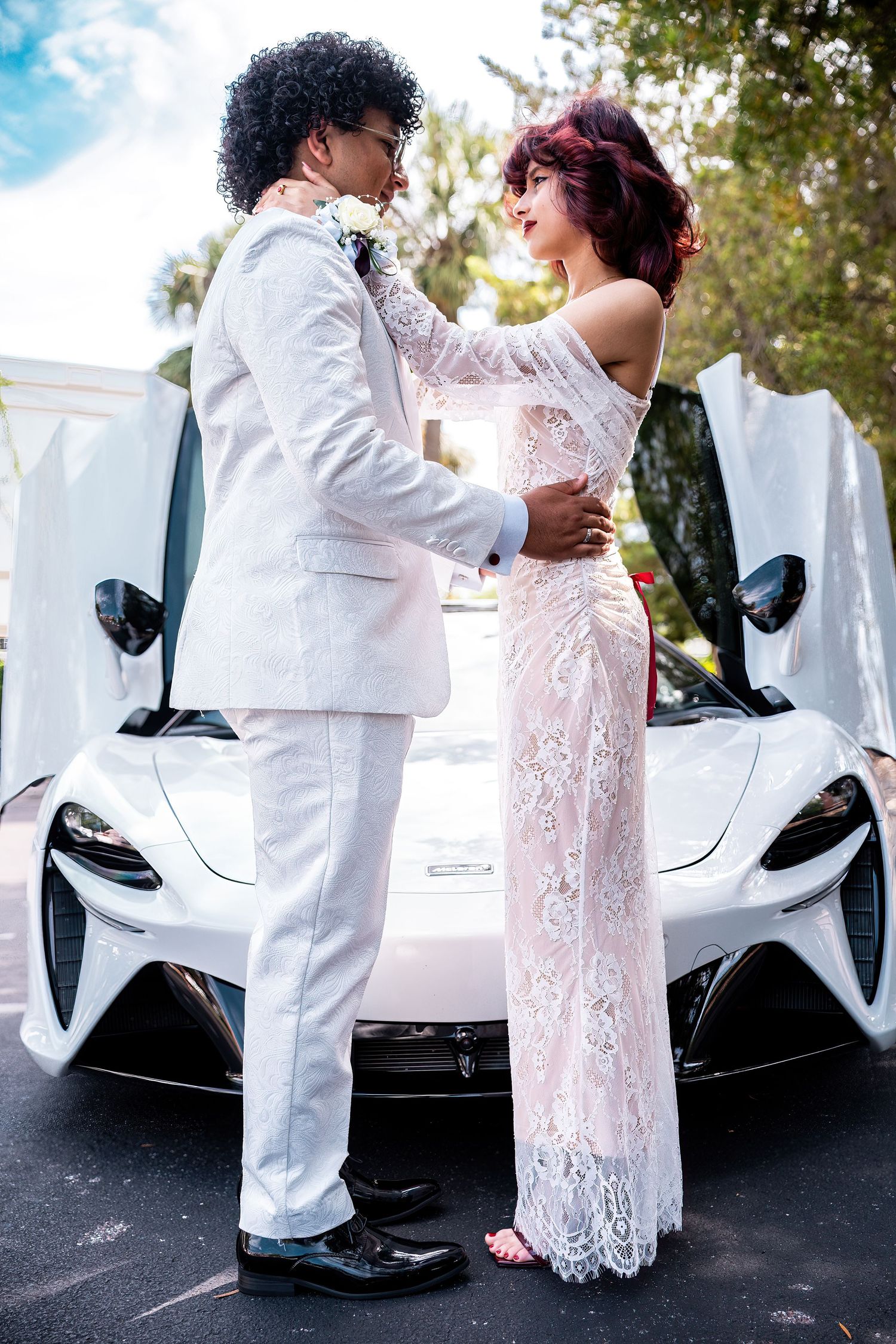 A couple in white formal attire embrace in front of a sleek McLaren sports car on their prom day.