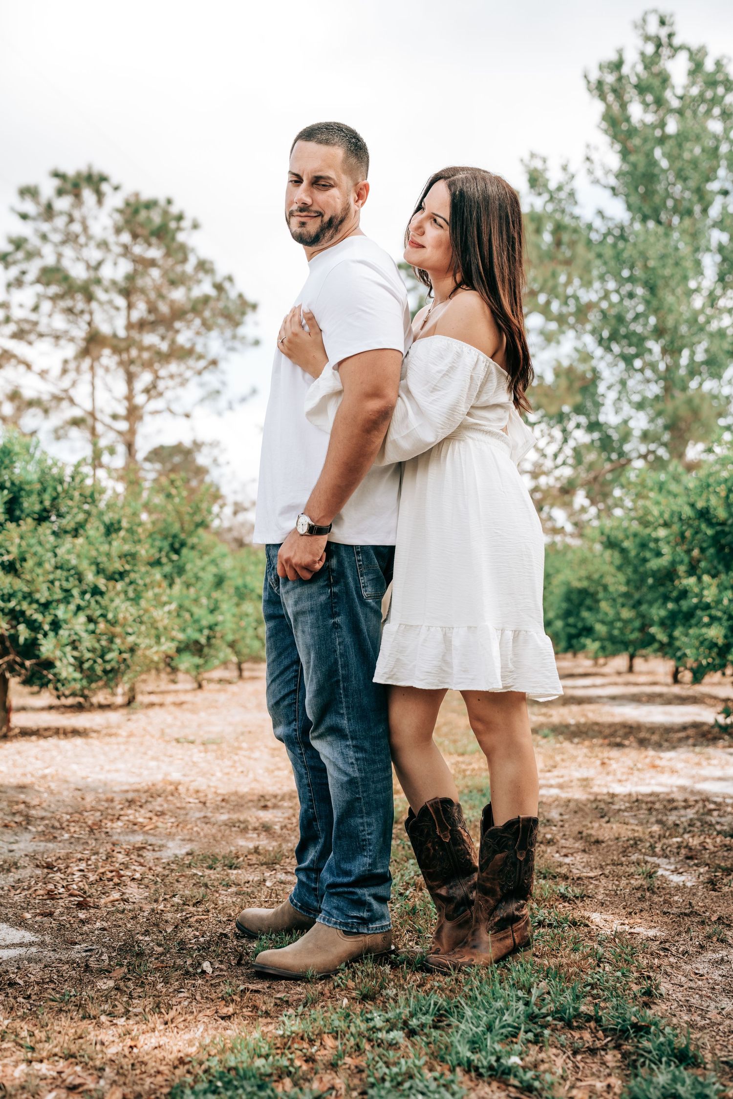 A couple in casual attire and cowboy boots stand together outdoors in a natural setting with trees and cloudy sky in the background.