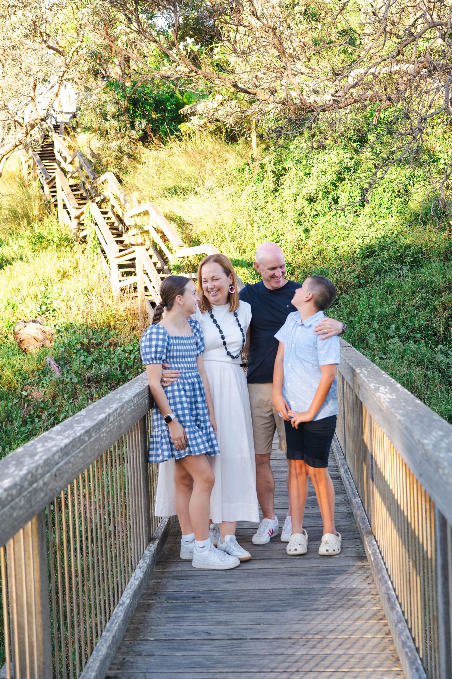 Family of four smiling together on a wooden bridge surrounded by lush greenery on a sunny day.