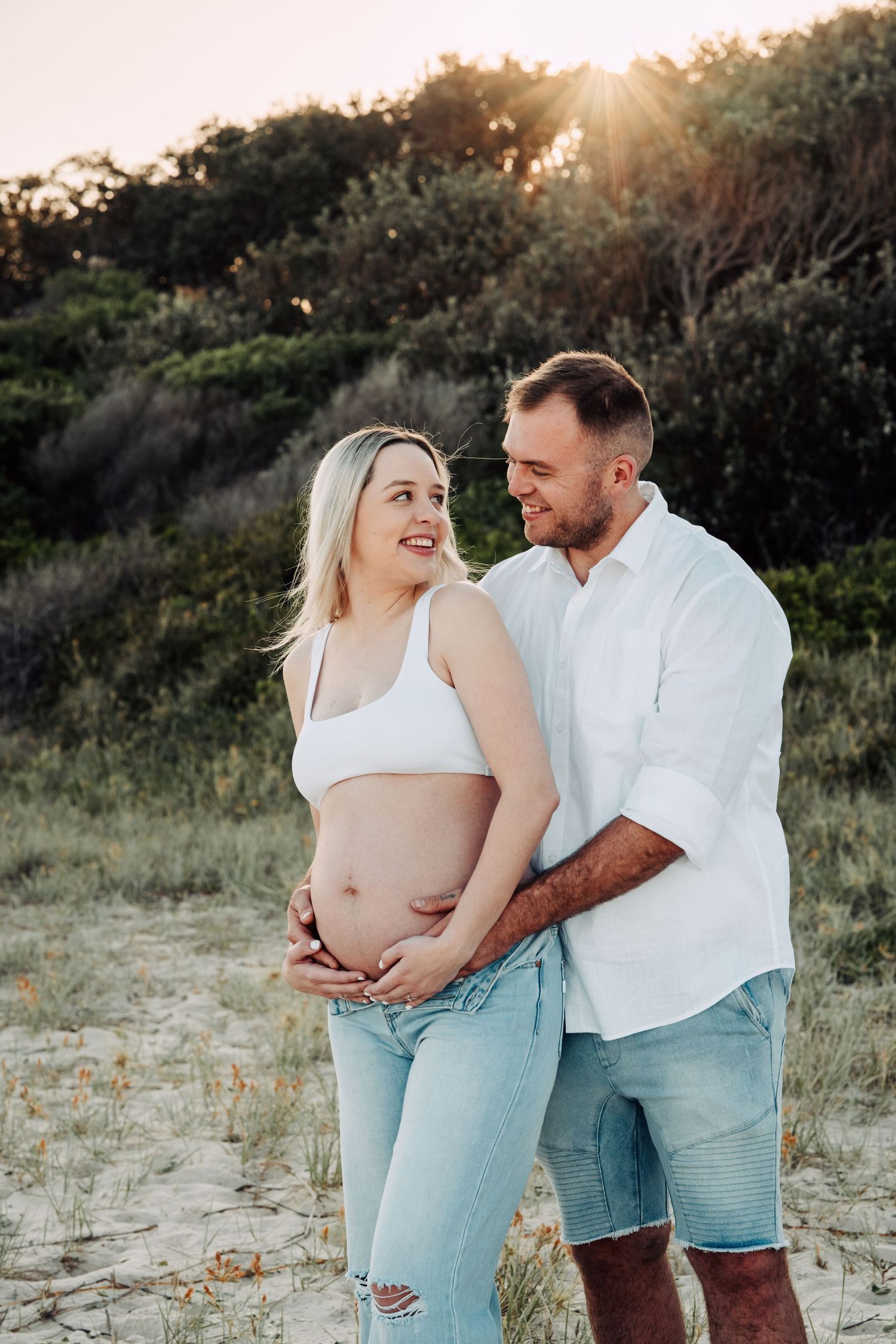 Pregnant couple embracing outdoors at sunset, woman in white crop top and jeans, man in white shirt, sandy dunes background.