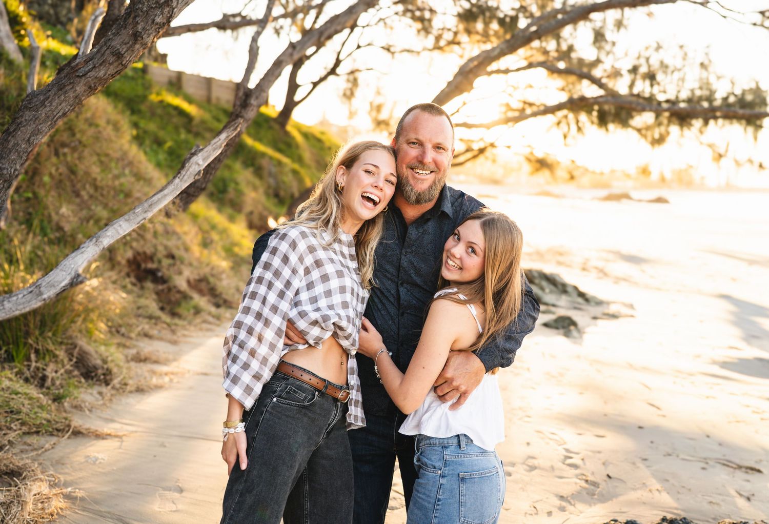 Family of three smiling and embracing on a sunny beach with trees and golden light in the background.