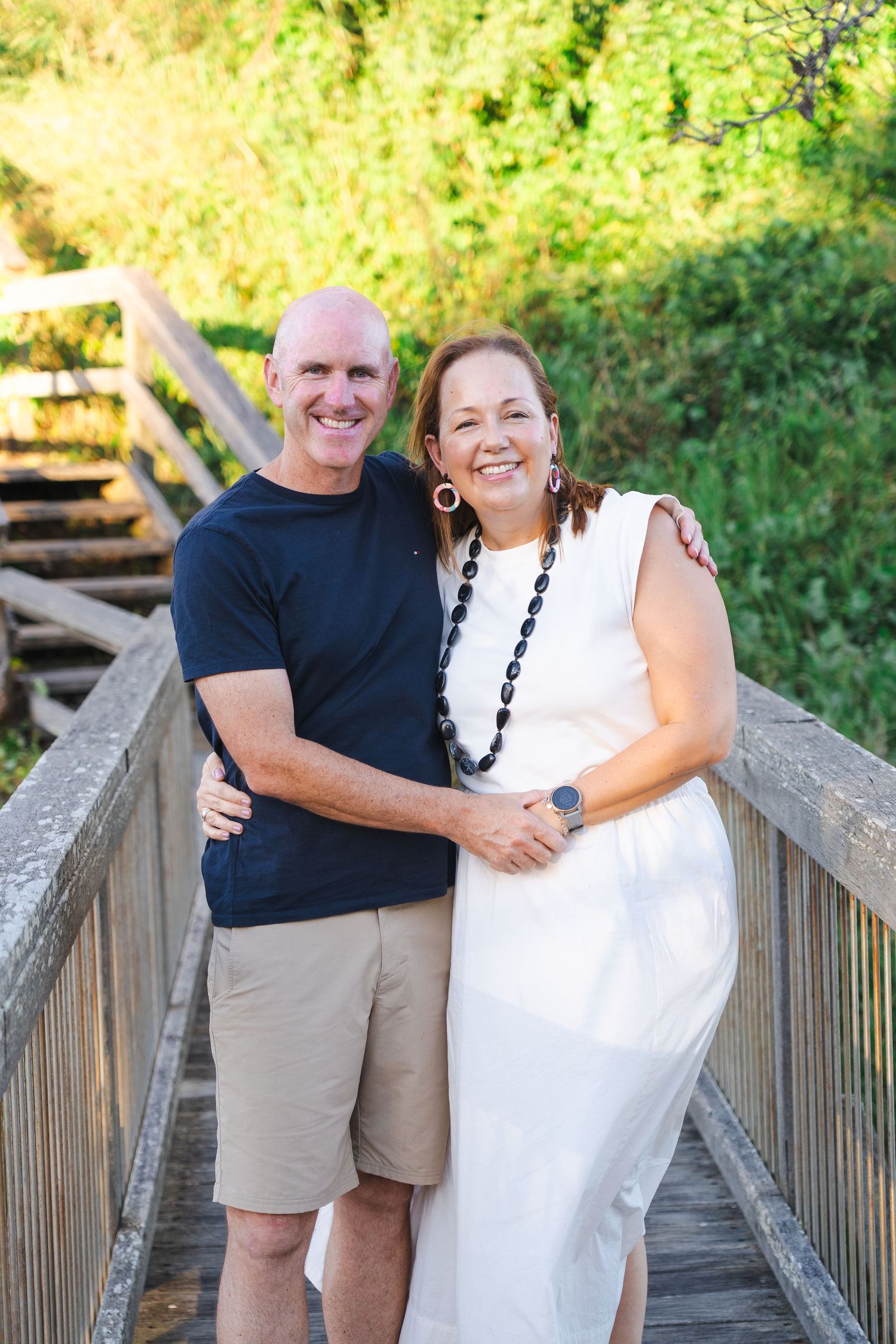 Smiling middle-aged couple embracing on a wooden bridge surrounded by lush greenery, wearing casual summer outfits.