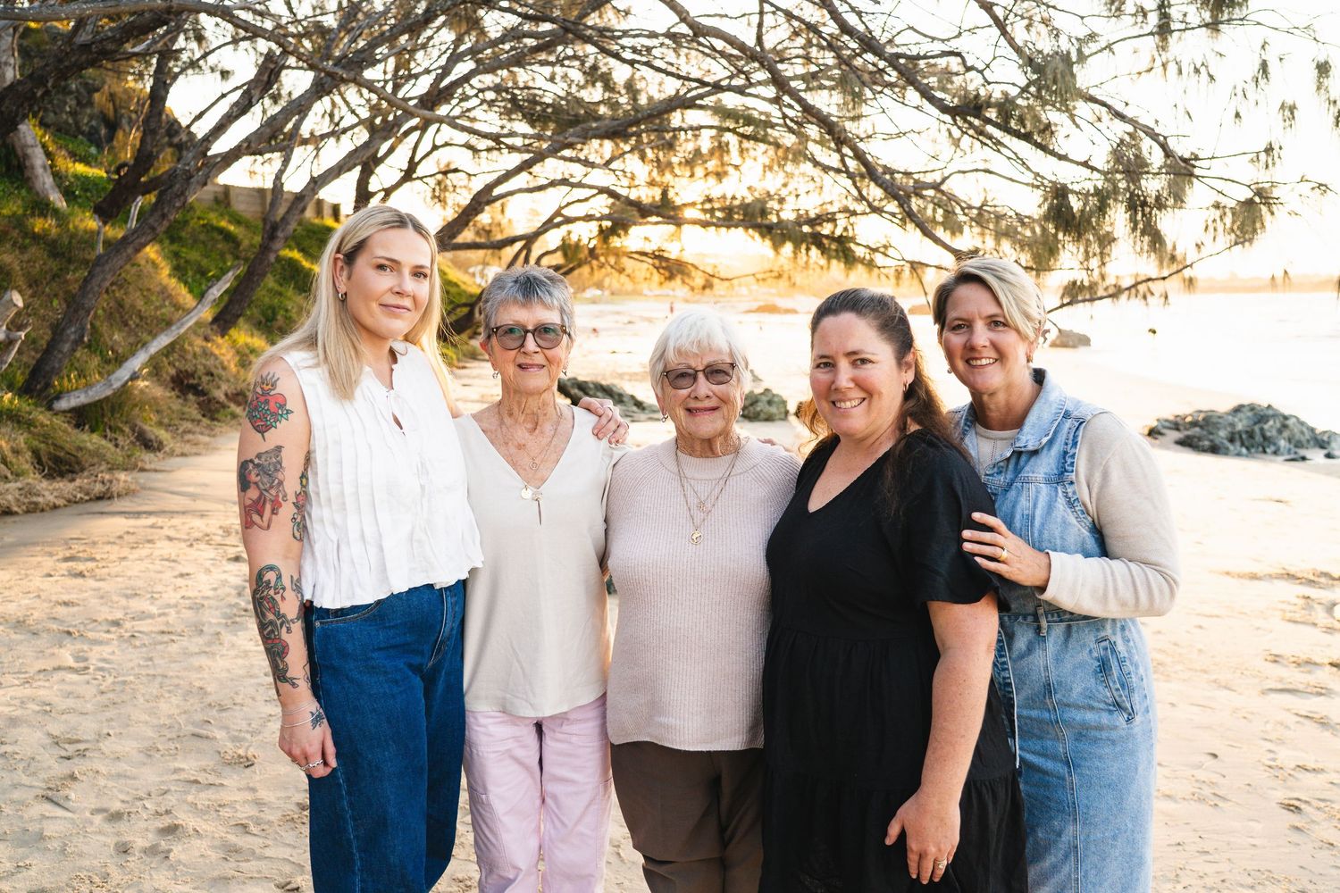 Five women smile together on a sandy beach beneath a tree at golden hour.