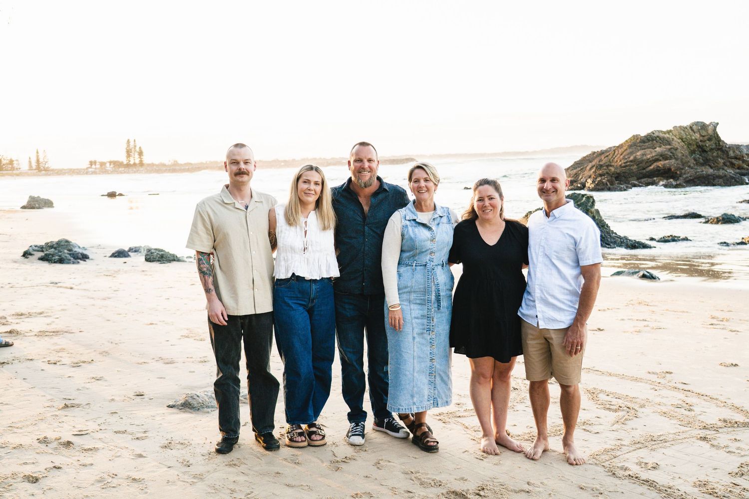 Six people smiling together on a sandy beach at sunset with rocky coastline in the background.