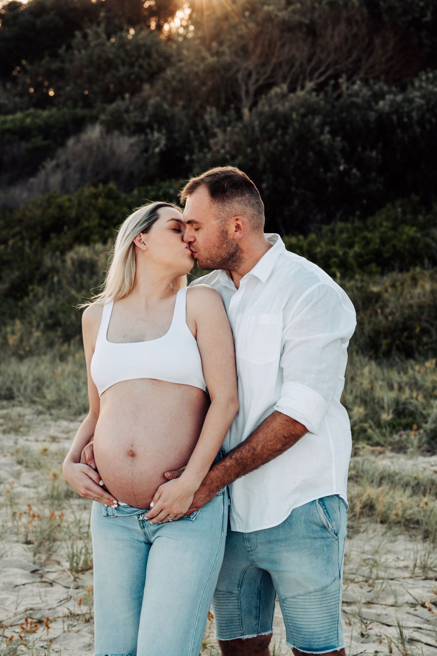 Couple sharing a kiss outdoors at sunset, man embracing pregnant woman's baby bump, wearing white tops and jeans.