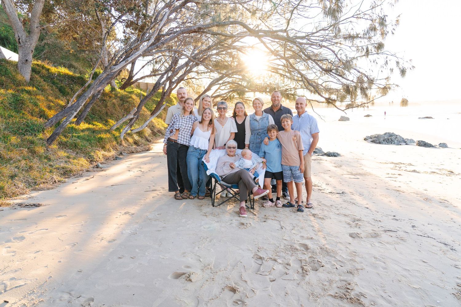 Family group photo on sandy beach at golden hour under trees with ocean backdrop.