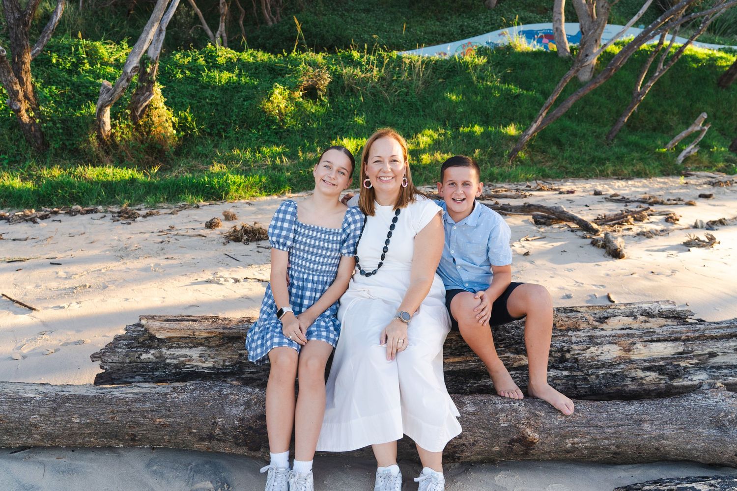 Woman and two children smiling on driftwood at sandy beach with tropical greenery in background.