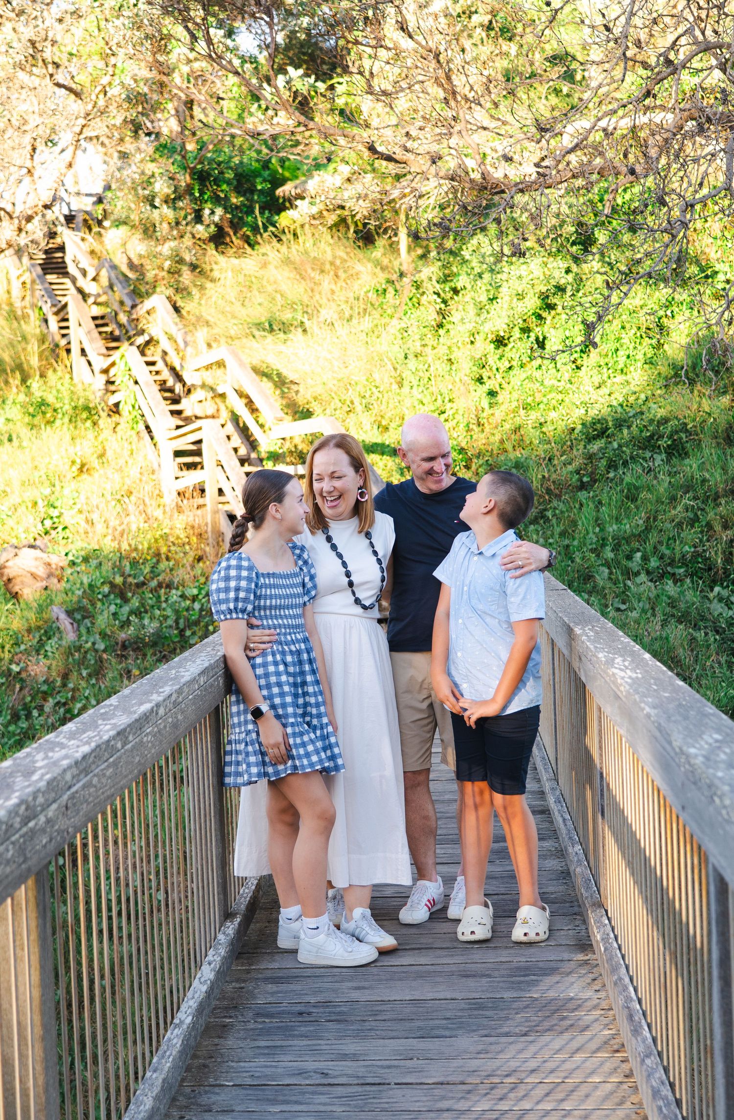 Family of four laughing together on a wooden bridge surrounded by lush greenery on a sunny day.