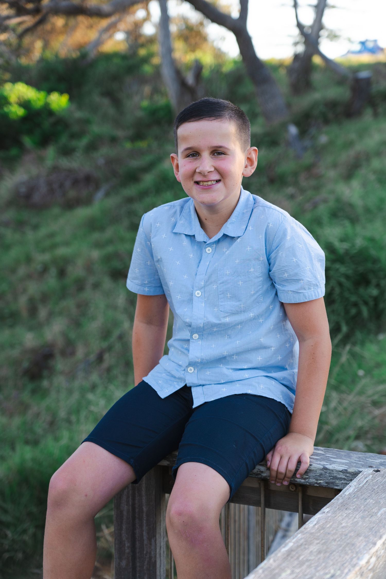 Smiling preteen boy in light blue shirt and navy shorts sitting on wooden fence outdoors with green hillside background.