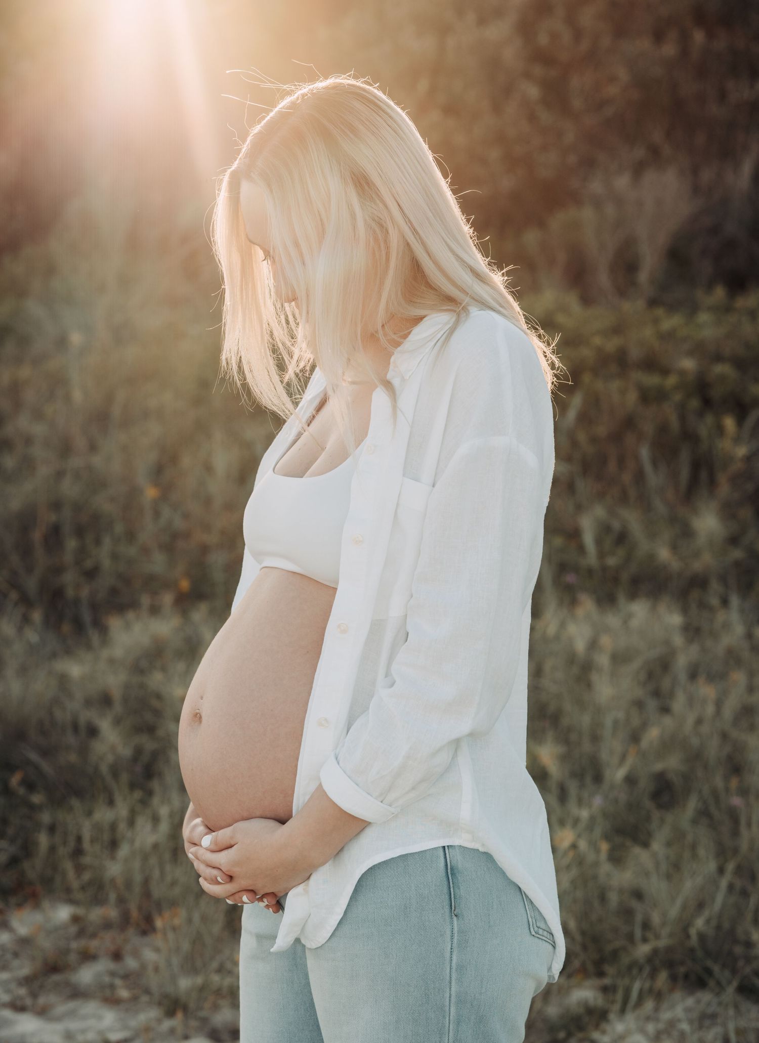Pregnant woman with blonde hair holding her baby bump outdoors in golden sunlight, wearing white shirt and jeans.