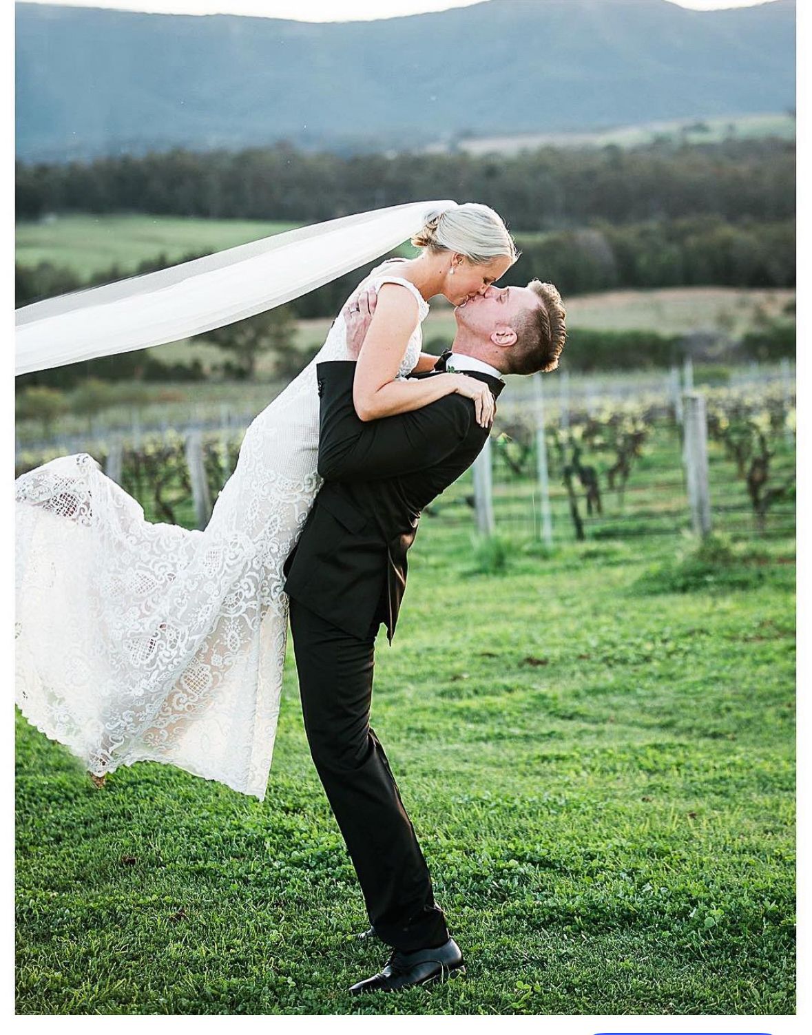 A romantic dip pose between a couple in formal wedding attire on a grassy hillside with scenic views.