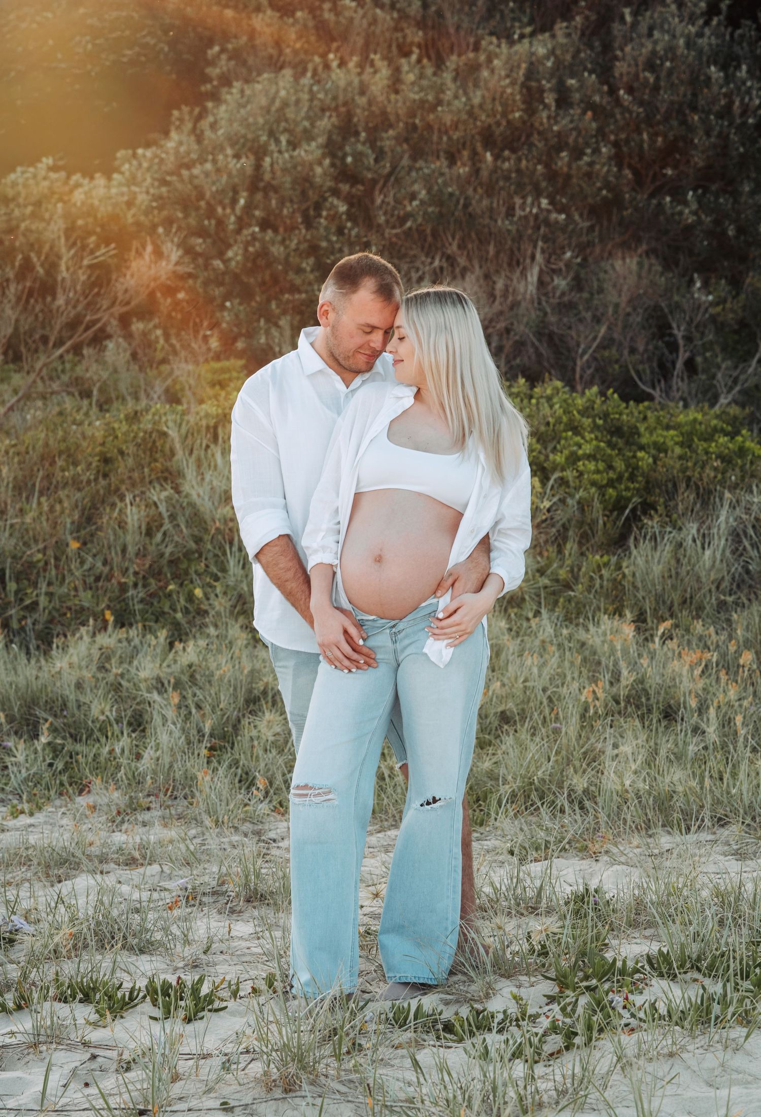 Couple in white shirts and jeans embrace on sandy beach during golden hour sunset.