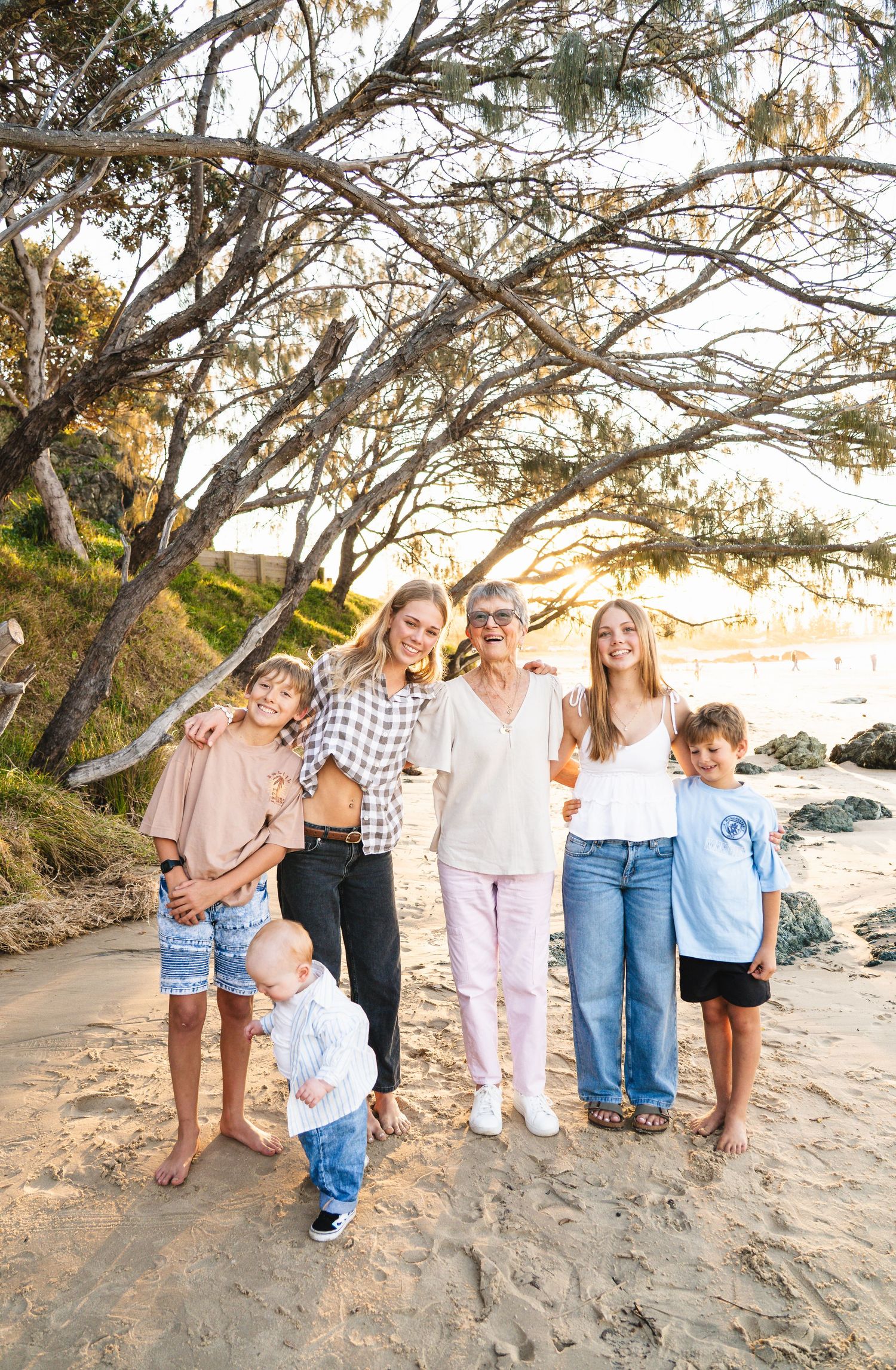 Family group of six posing on sandy beach under trees at golden sunset hour.