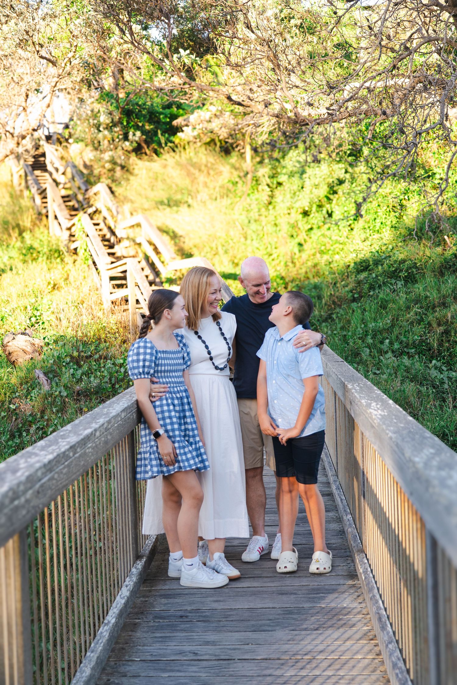 Family of four smiling together on a wooden boardwalk surrounded by lush greenery on a sunny day.